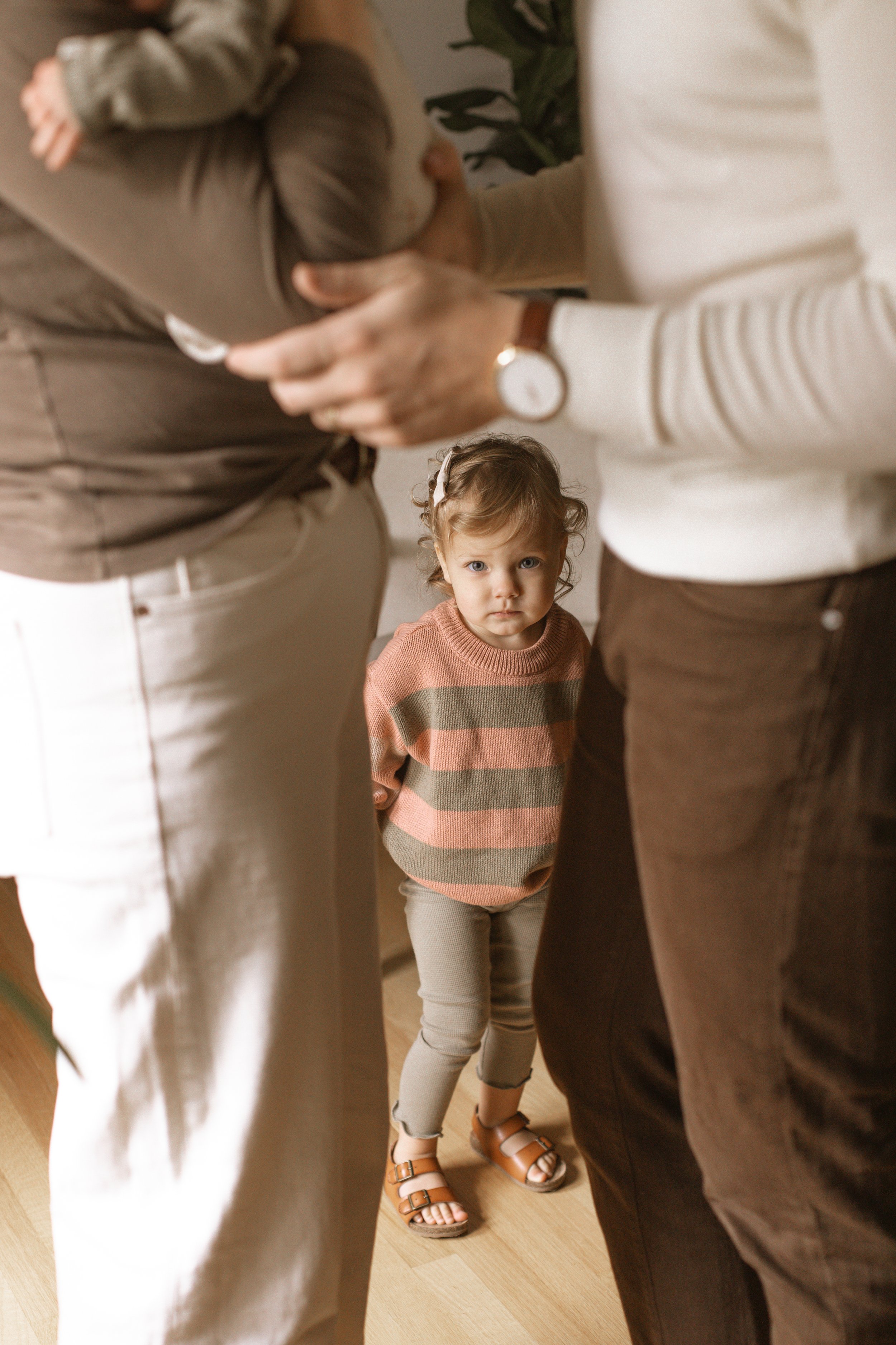 A young girl with curly hair and a serious expression stands between two adults, looking directly at the camera, while the adults are engaged in a conversation. Big sister in a newborn session. Newborn Lifestyle Photographer serving Evanston, Chicago
