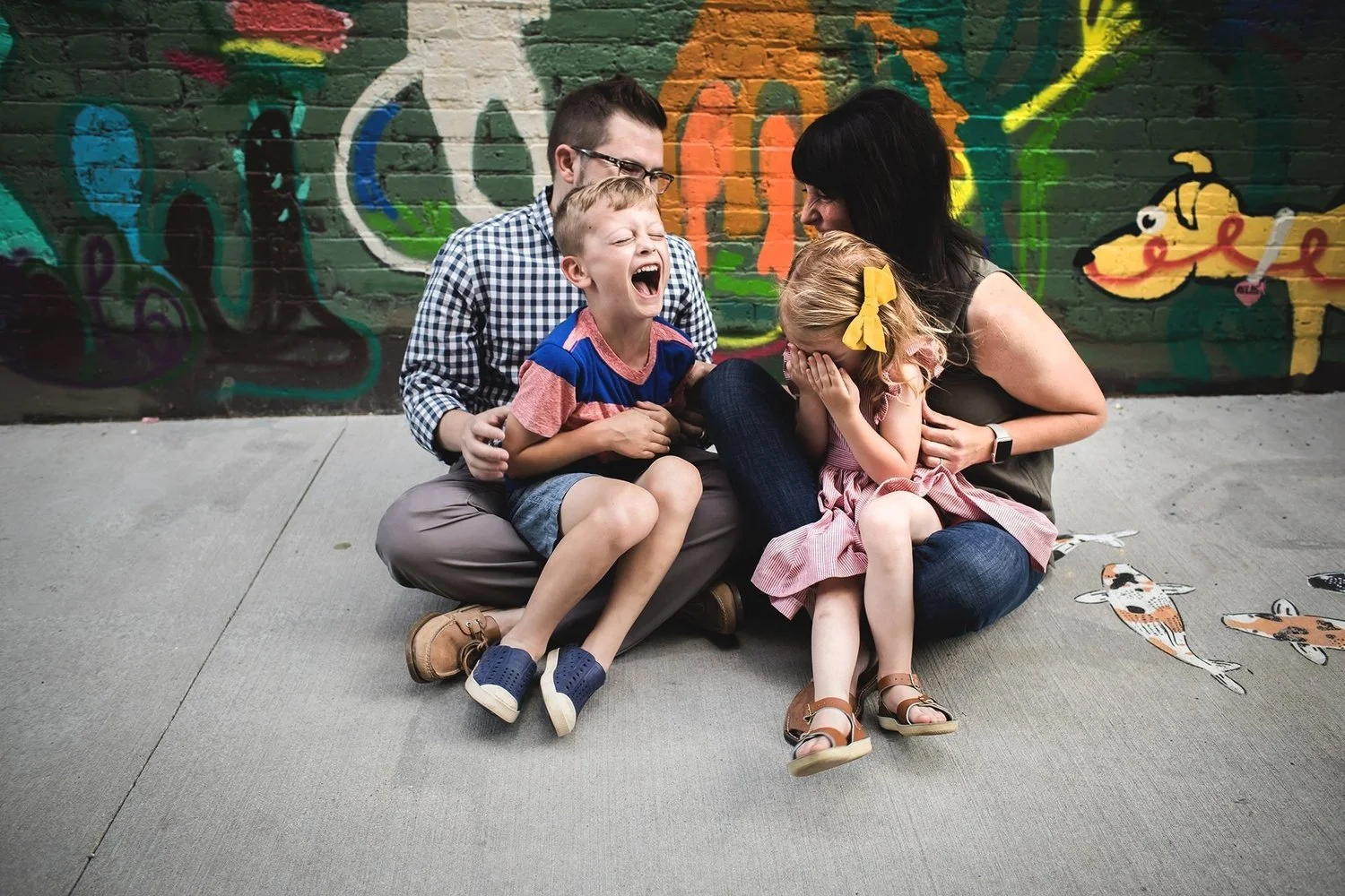 A family of four, with a mother, father, young boy, and young girl, sitting on the sidewalk against a colorful graffiti wall. The boy is laughing with his eyes closed, and the girl is covering her face with her hands, smiling. The parents are engagin