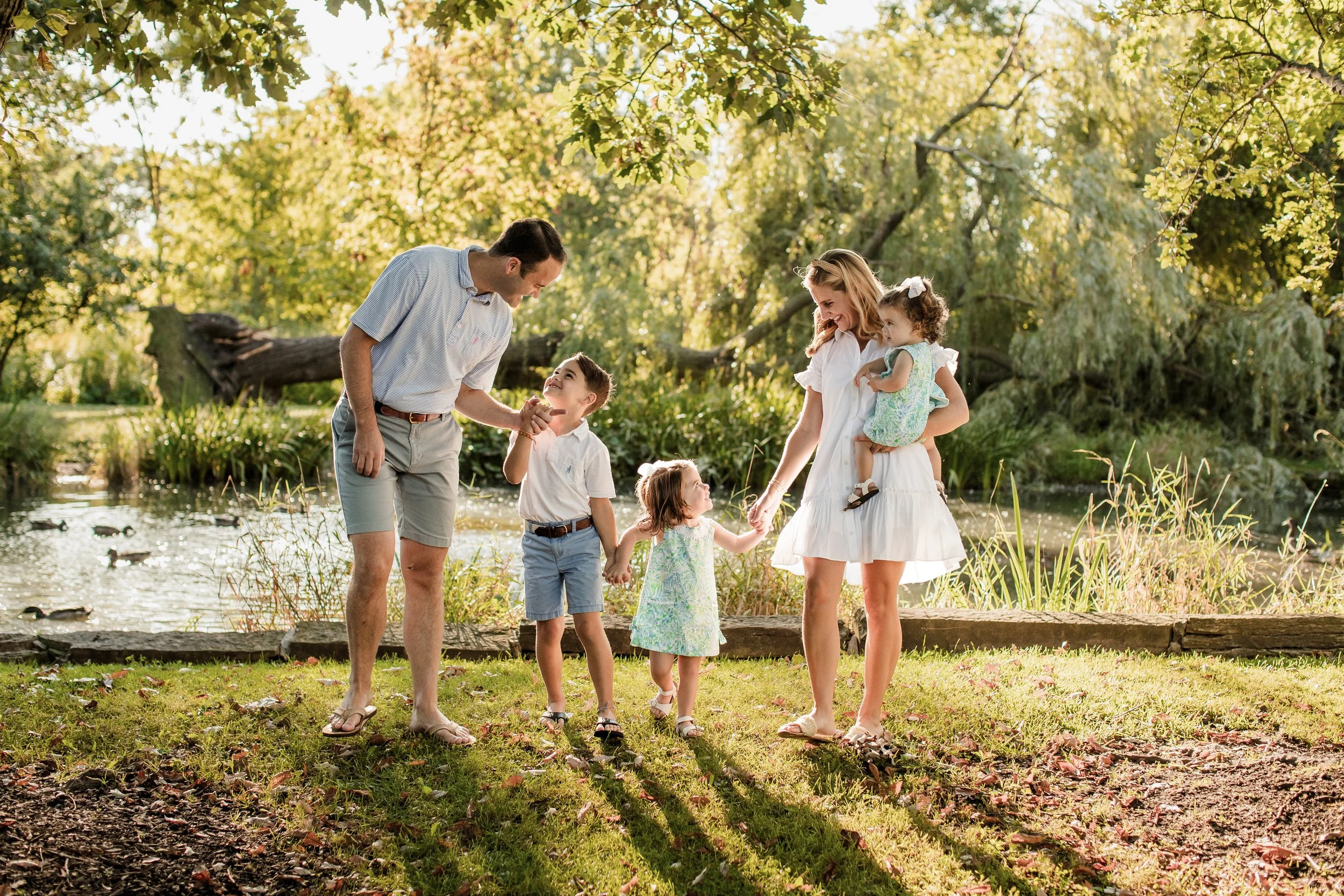 A family of six enjoying a sunny day by a pond in a park, with trees and greenery in the background. The father is holding hands with a girl, while the mother holds a toddler in her arms. A young boy is smiling and standing next to the father. The fa
