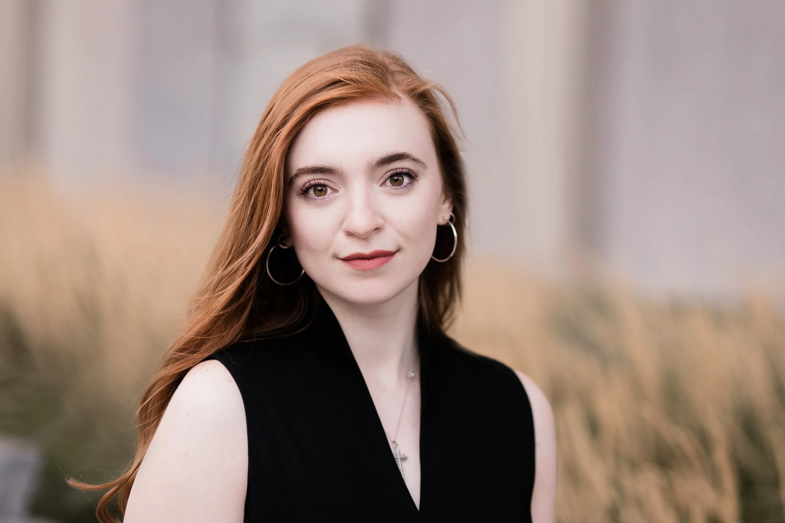Portrait of a young woman with red hair, wearing a black sleeveless top, hoop earrings, and a silver necklace, outdoors with a blurred background.