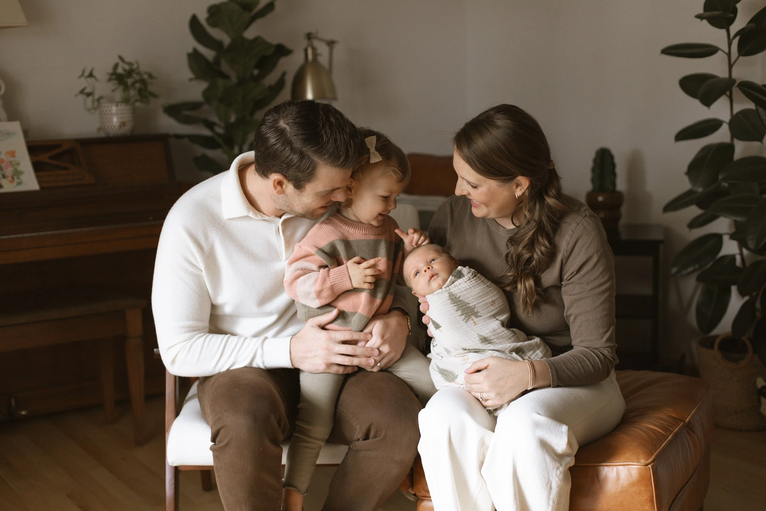A family of four sitting together indoors, smiling and looking at their newborn baby wrapped in a blanket, with a young girl touching the baby's head, in a cozy living room. Lifestyle newborn session shot in Evanston IL.