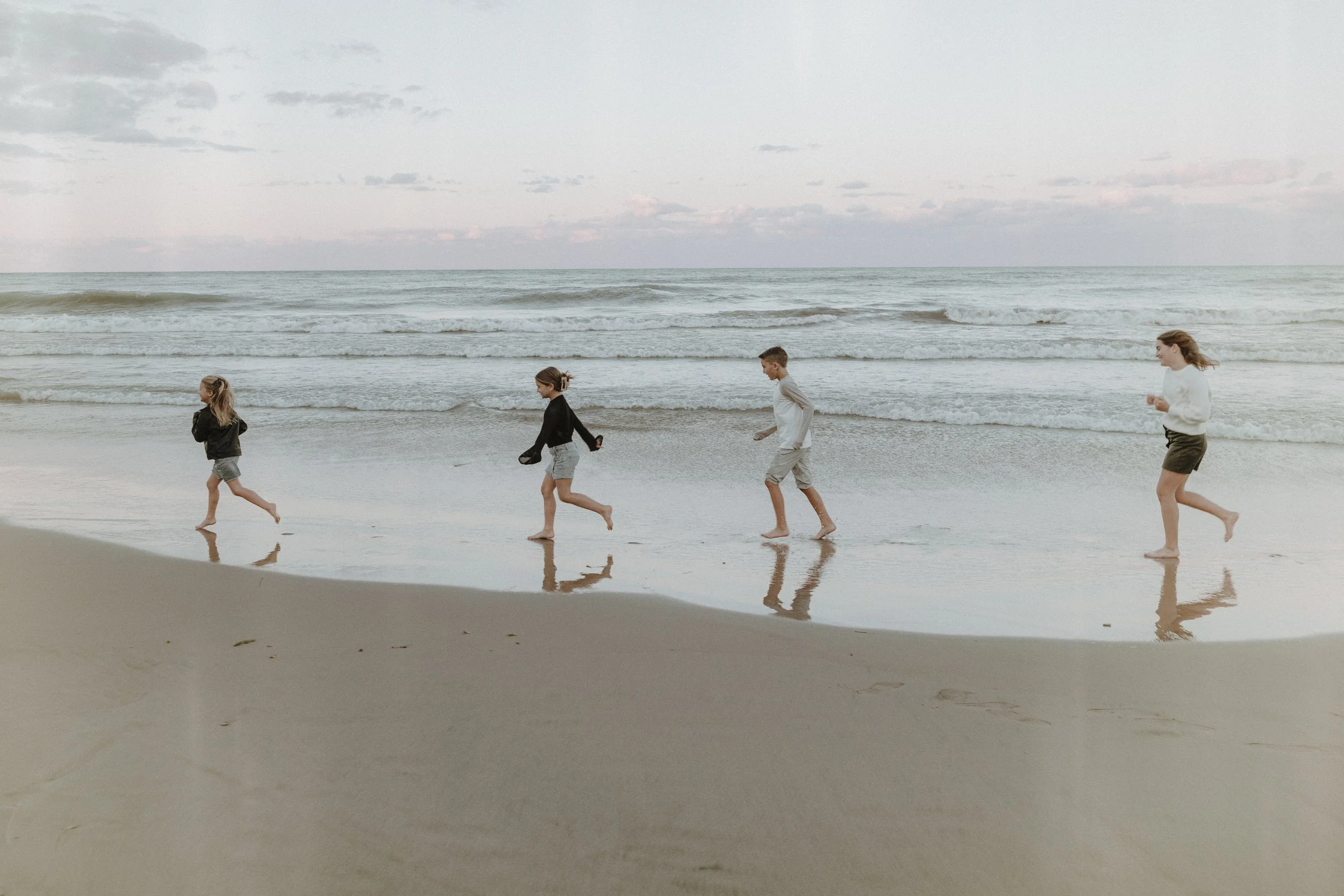 Four children walking along the shoreline of a beach during early morning or evening, with gentle waves and a cloudy sky in the background. Lifestyle Family Photography Session Evanston, IL.