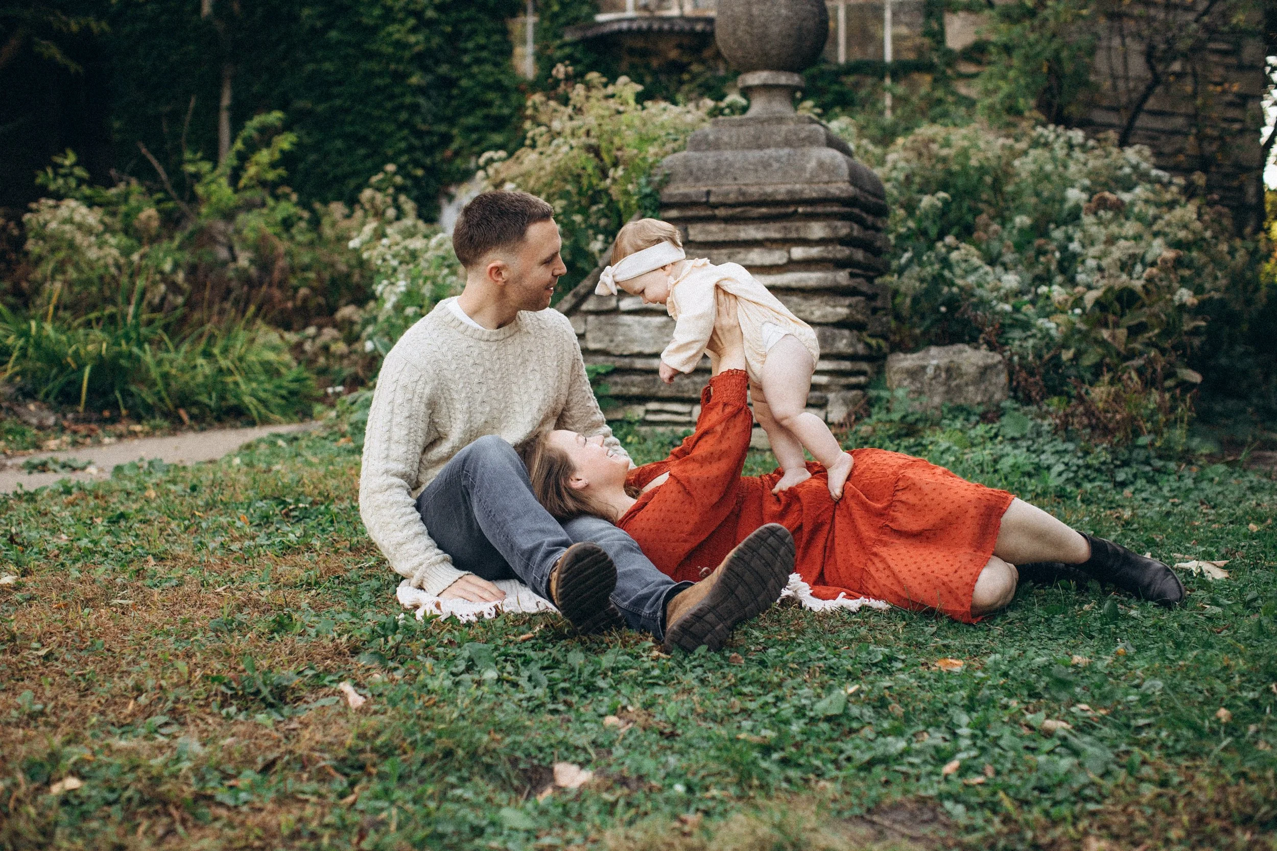 Family of three playing on the grass outside, with a man, woman, and baby girl. The woman lies on her back holding the baby above her. The man sits nearby, all smiling.