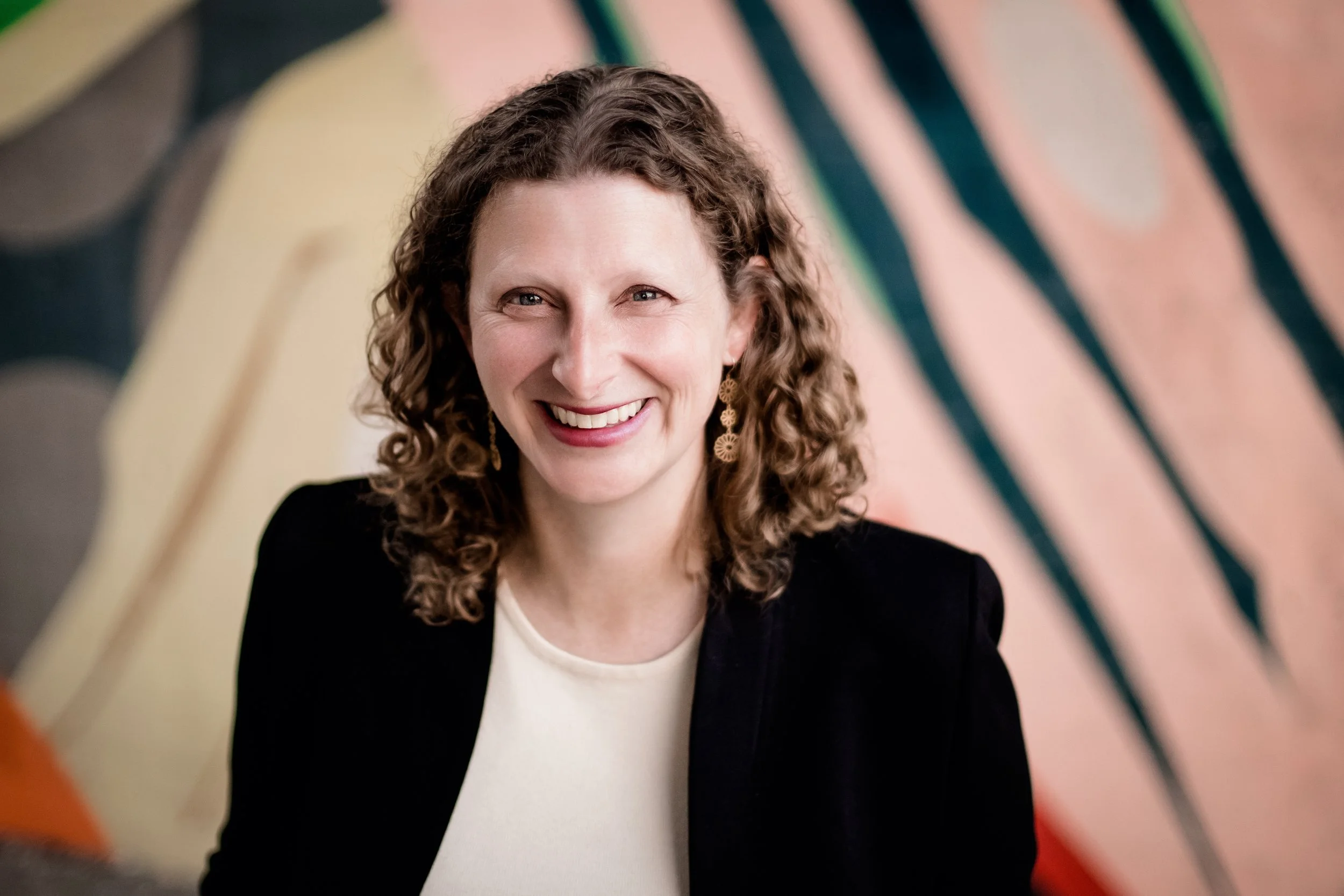 A woman with curly hair smiling, wearing earrings and a black jacket, in front of a colorful abstract background.
