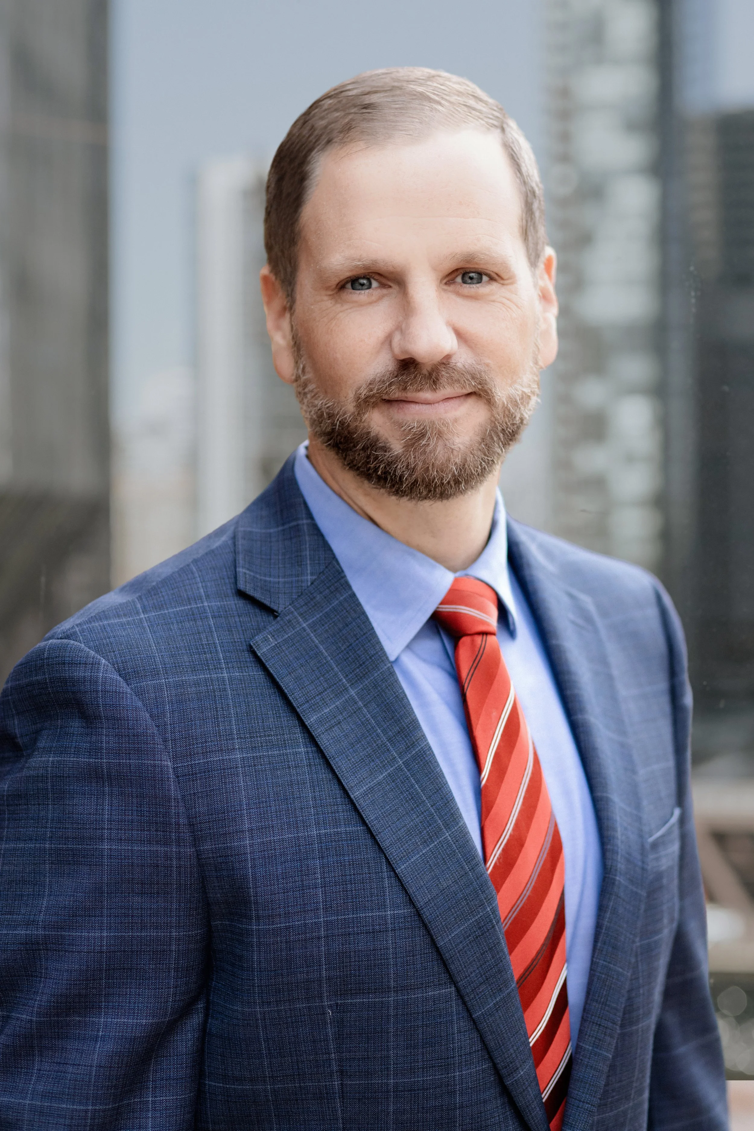 A middle-aged man with a beard and short hair wearing a blue checked suit, light blue shirt, and red striped tie, standing in front of a window with a cityscape background.