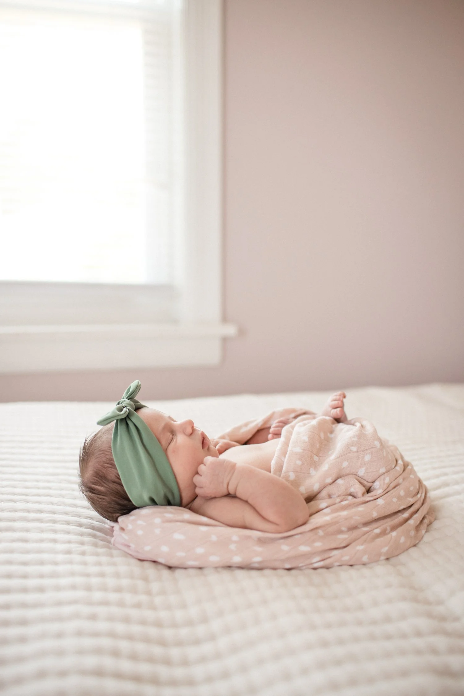 A sleeping baby girl lying on a bed, wrapped in a pink polka dot blanket, wearing a green headband with a bow, in a room with soft natural light from a nearby window. Newborn Photography in Chicago IL