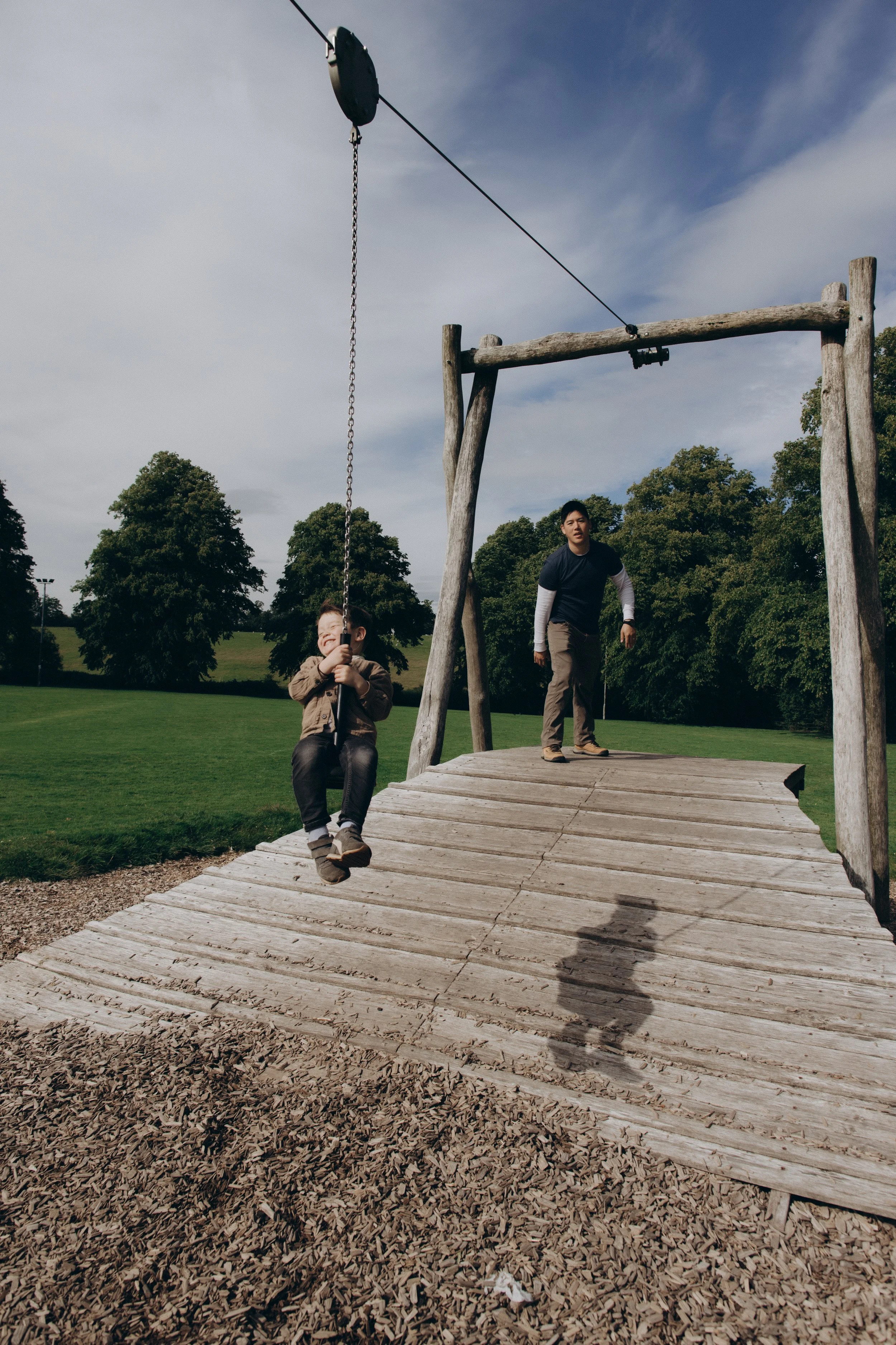 A young boy is swinging on a playground swing, smiling, while an adult man is standing on a wooden platform nearby, in an open park with trees in the background under a partly cloudy sky.