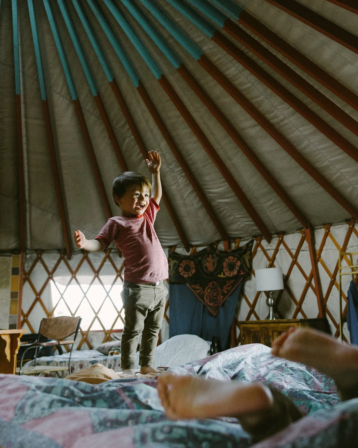 A young boy standing on a bed inside a yurt, smiling and raising his right hand. The interior features wooden lattice walls, a window with curtains, a small table, and a lamp.