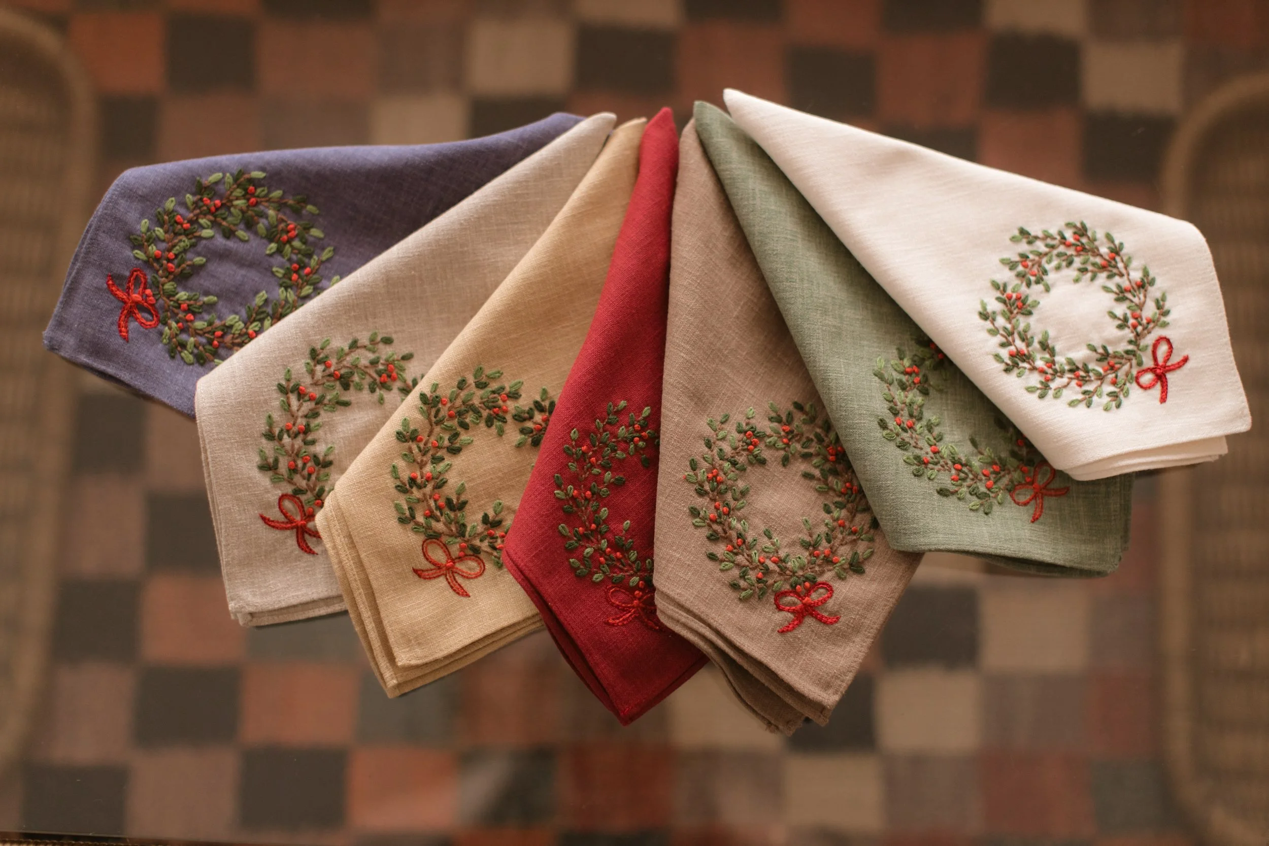 Five folded cloth napkins with embroidered wreath designs and red bows, in purple, beige, red, green, and white colors, arranged on a transparent surface. Product Photography Shoot Evanston, IL.
