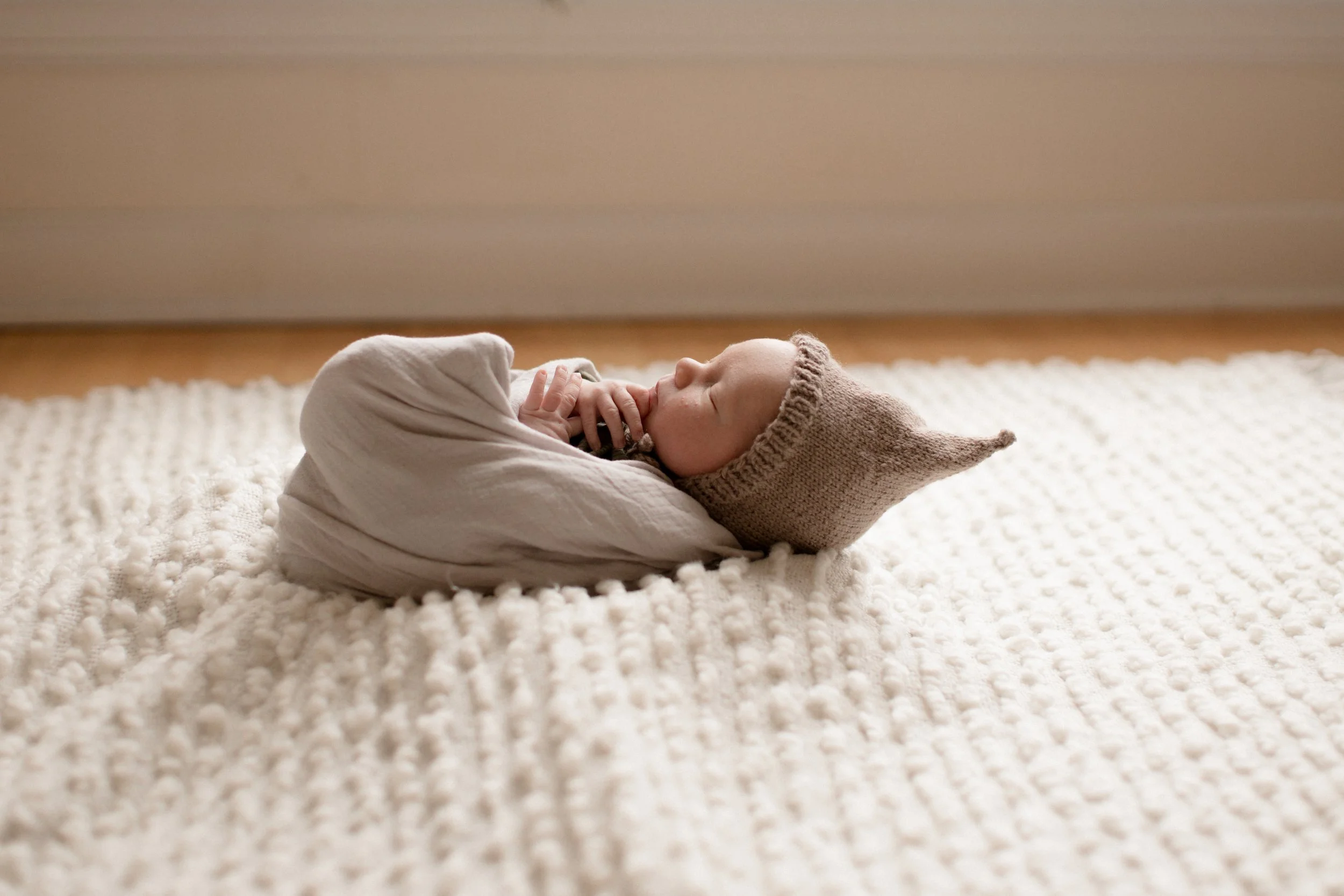 A sleeping baby wrapped in a blanket, wearing a knitted hat with a pointy tip, lying on a textured cream-colored rug. Lifestyle newborn session shot in Chicago IL.