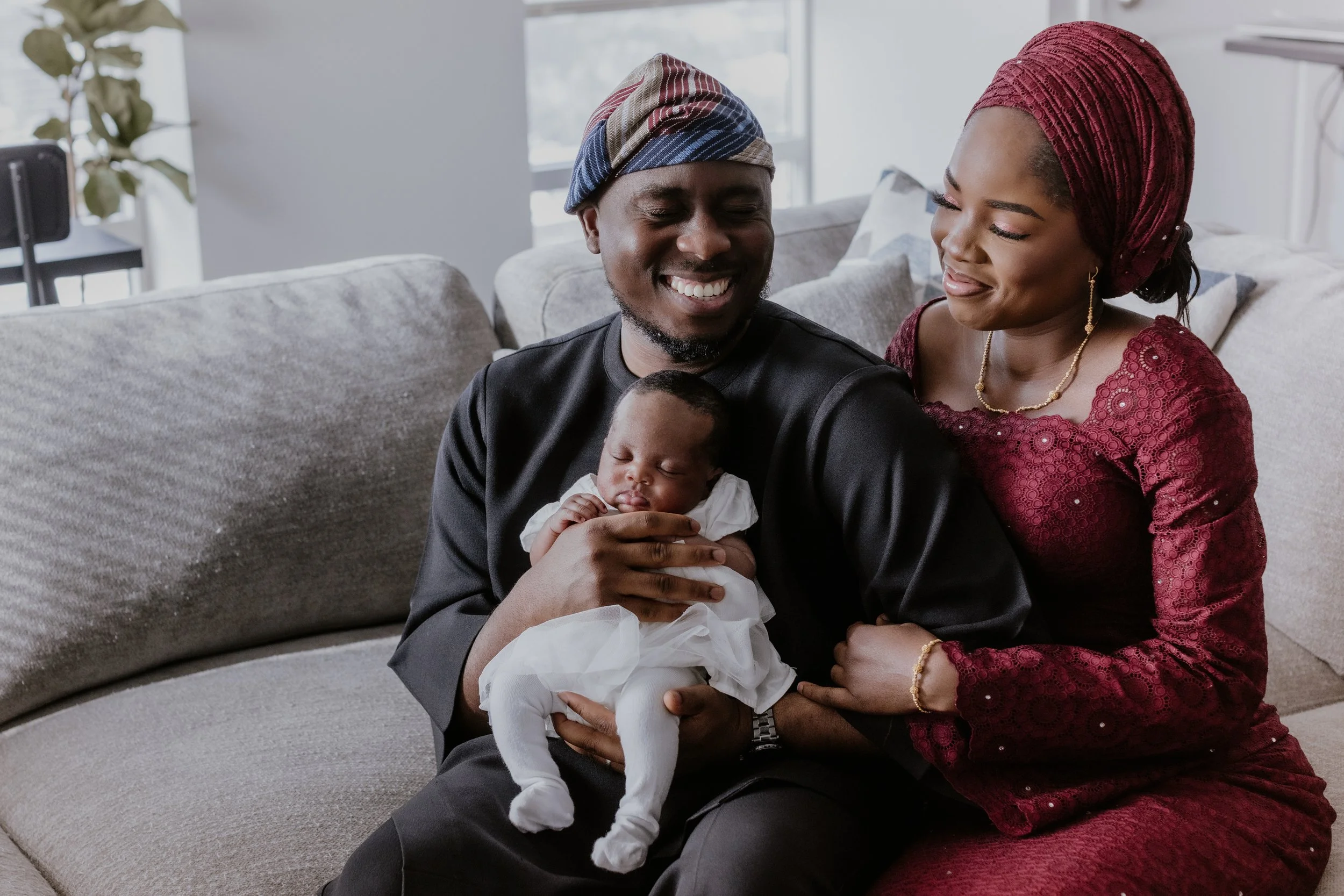 A smiling African-American family with a man holding a sleeping baby, a woman sitting next to him smiling, on a gray couch in a bright living room.