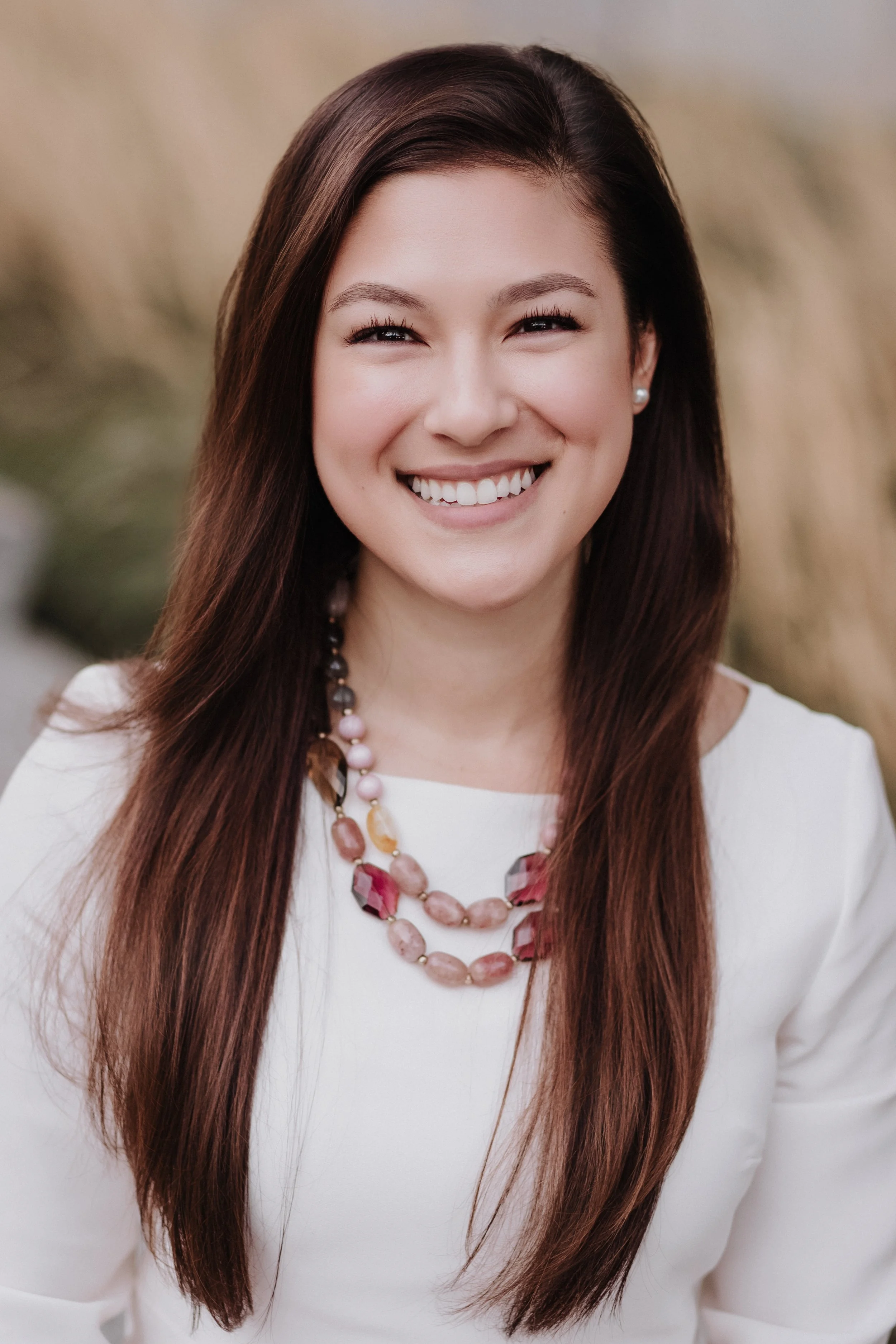 A woman with long brown hair and a broad smile, wearing a white blouse and a colorful beaded necklace, standing outdoors with a blurred natural background.