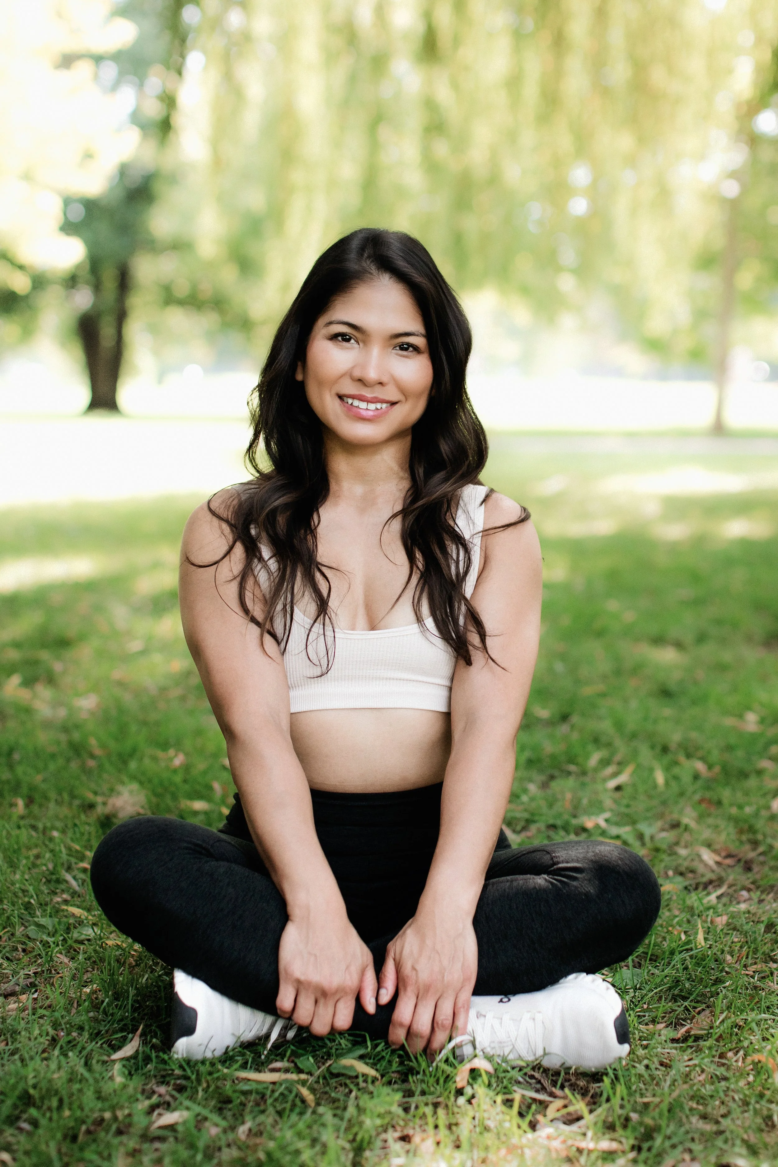 A woman sitting cross-legged on grass in a park, smiling at the camera, with trees and green foliage in the background.