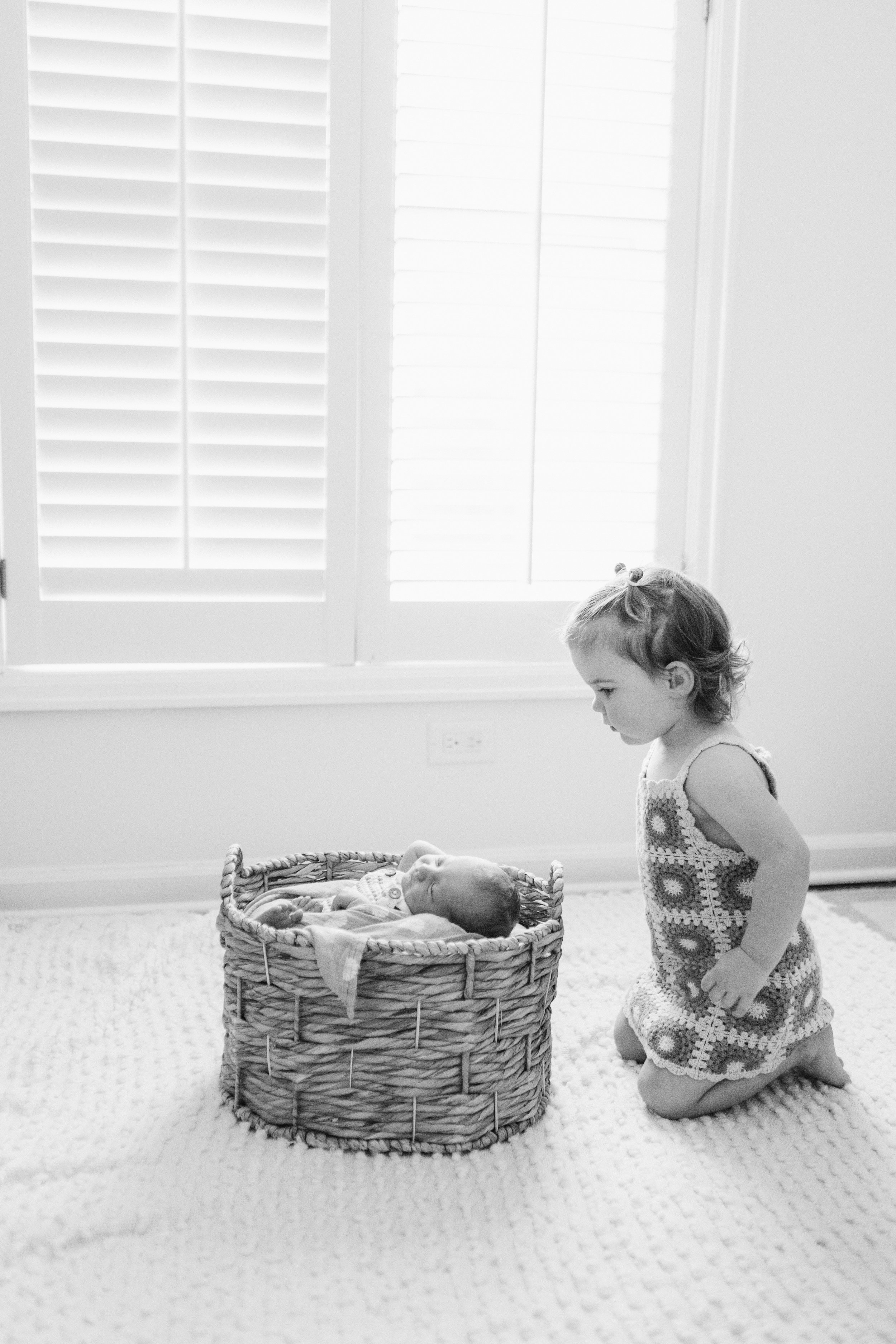 A young girl kneeling on a textured rug, looking at a baby lying in a woven basket. Lifestyle newborn session shot in Chicago IL