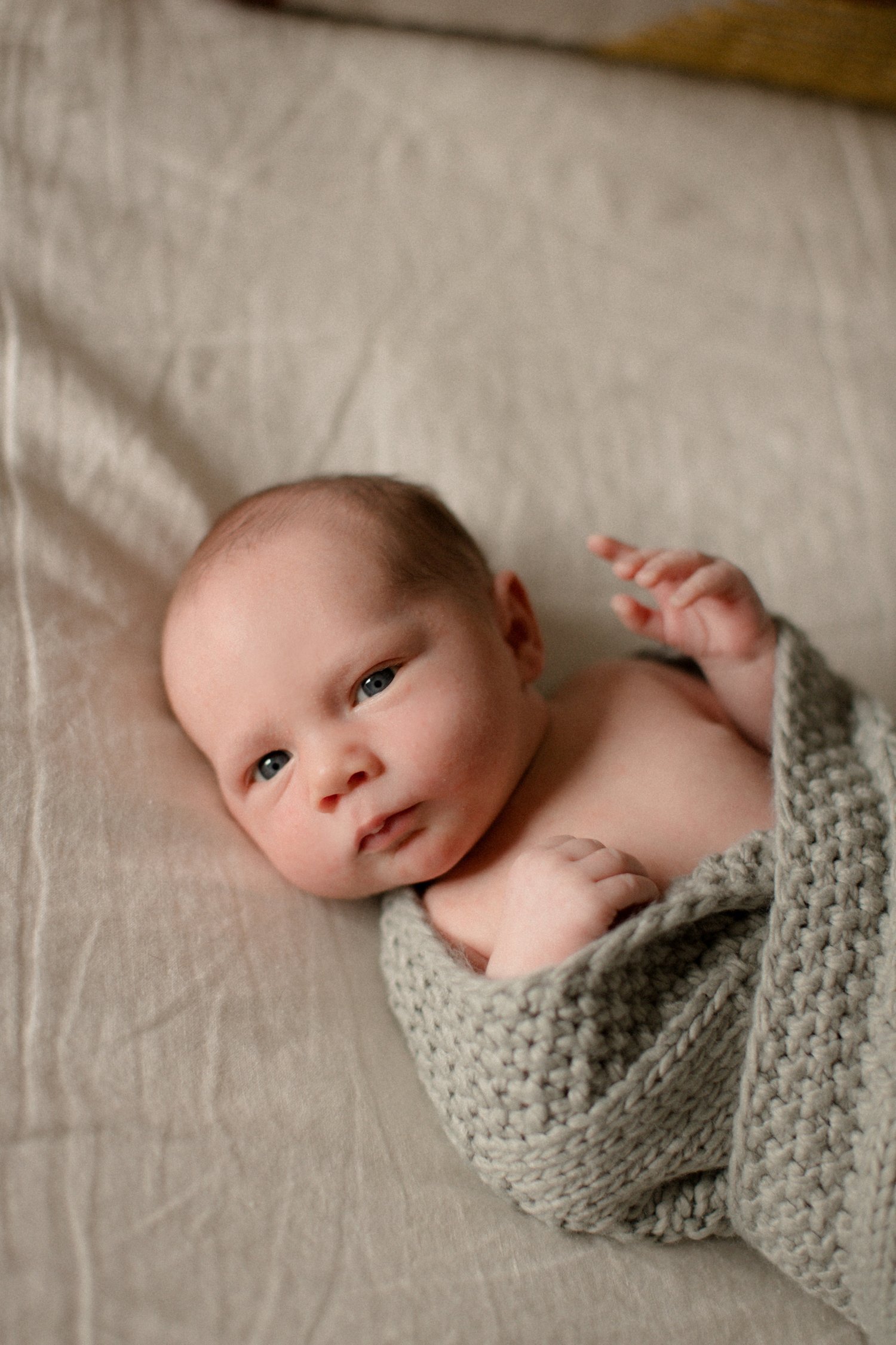 A baby lying on a beige surface, wrapped in a chunky gray knitted blanket, looking at the camera with blue eyes.  Lifestyle newborn photography session in Chicago IL