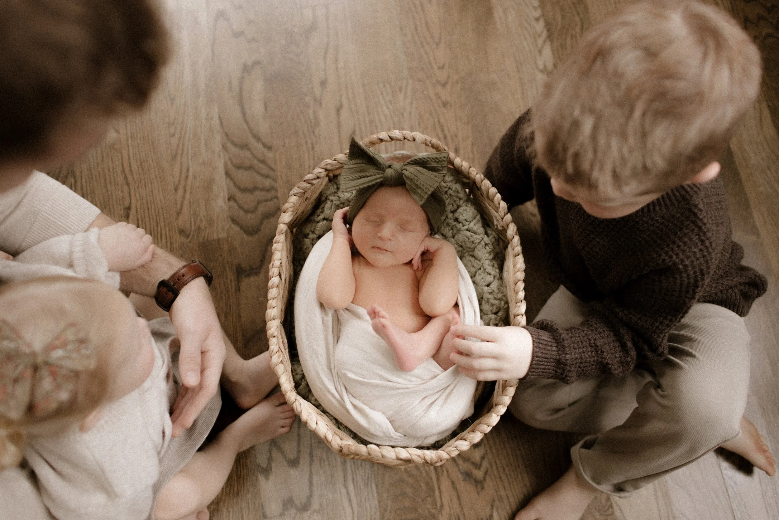 A newborn baby wrapped in a white blanket and lying in a woven basket, wearing a dark green bow headband, surrounded by two children and an adult, all sitting on a wooden floor.