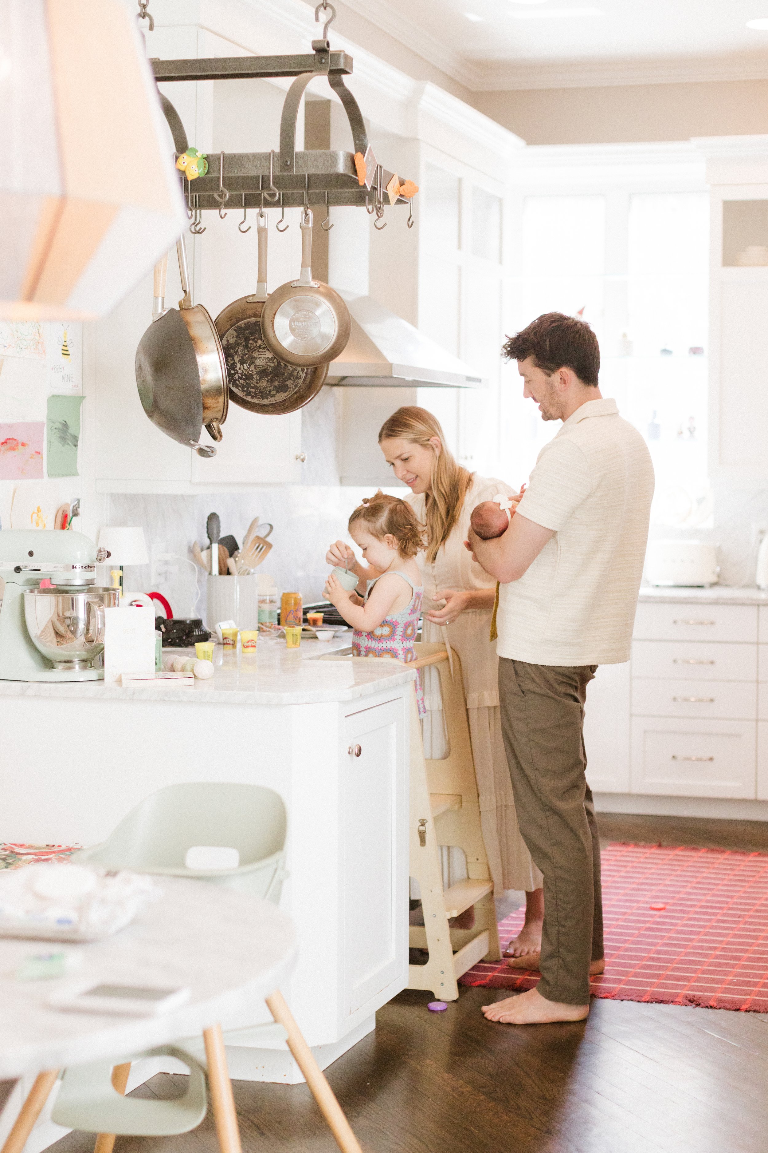Family in a bright white kitchen with pots hanging overhead, a little girl and her mother preparing food, father holding a baby nearby. Lifestyle newborn session shot in Chicago IL.