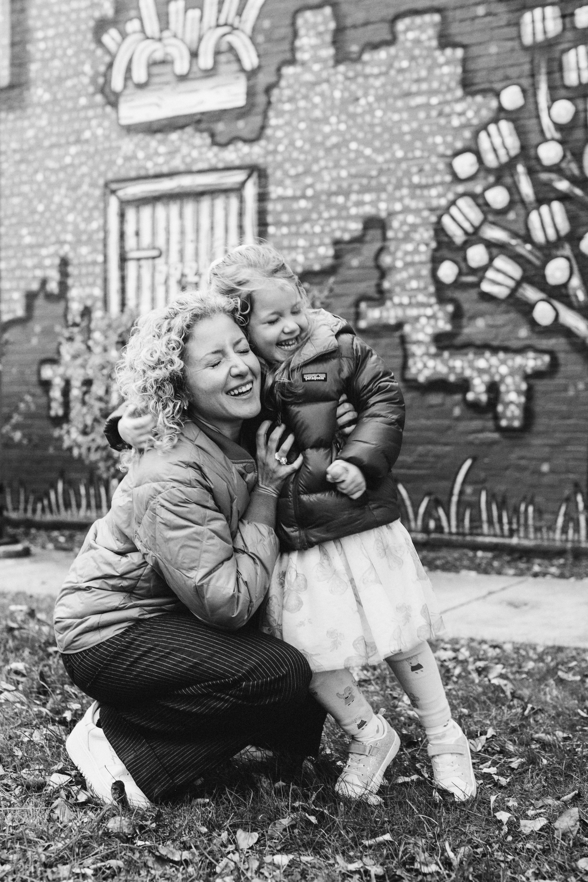 A woman and a young girl are sharing a joyful hug in front of a colorful, artistic mural on a brick wall. The woman is kneeling, laughing, and the girl is smiling, wearing a puffy jacket, a dress, and sneakers. The ground is covered with fallen leave