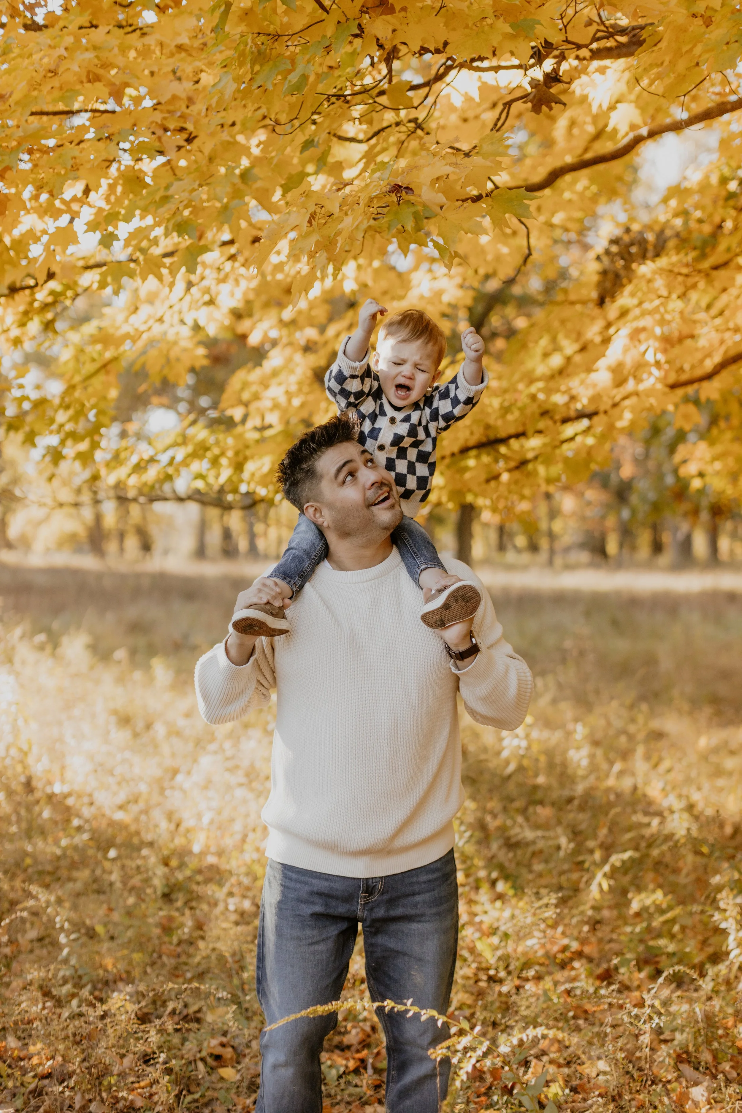 A man is giving a young boy a piggyback ride in a park with autumn-colored leaves. The boy is raising his fists and appears to be shouting or excited. The background is filled with yellow and orange fall foliage.
