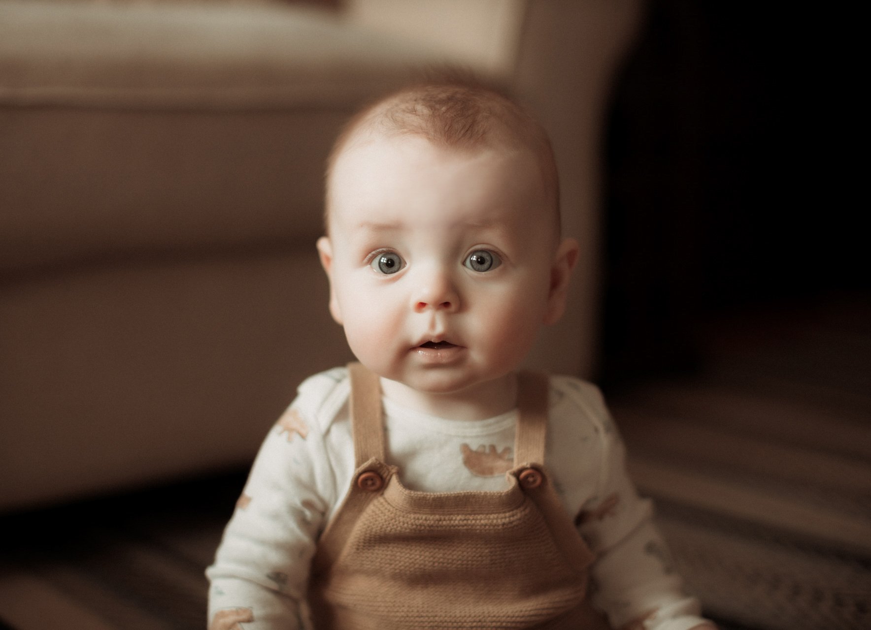 A young child with big blue eyes and short light brown hair, sitting on a striped blanket or bedspread, looking directly at the camera with a surprised expression.