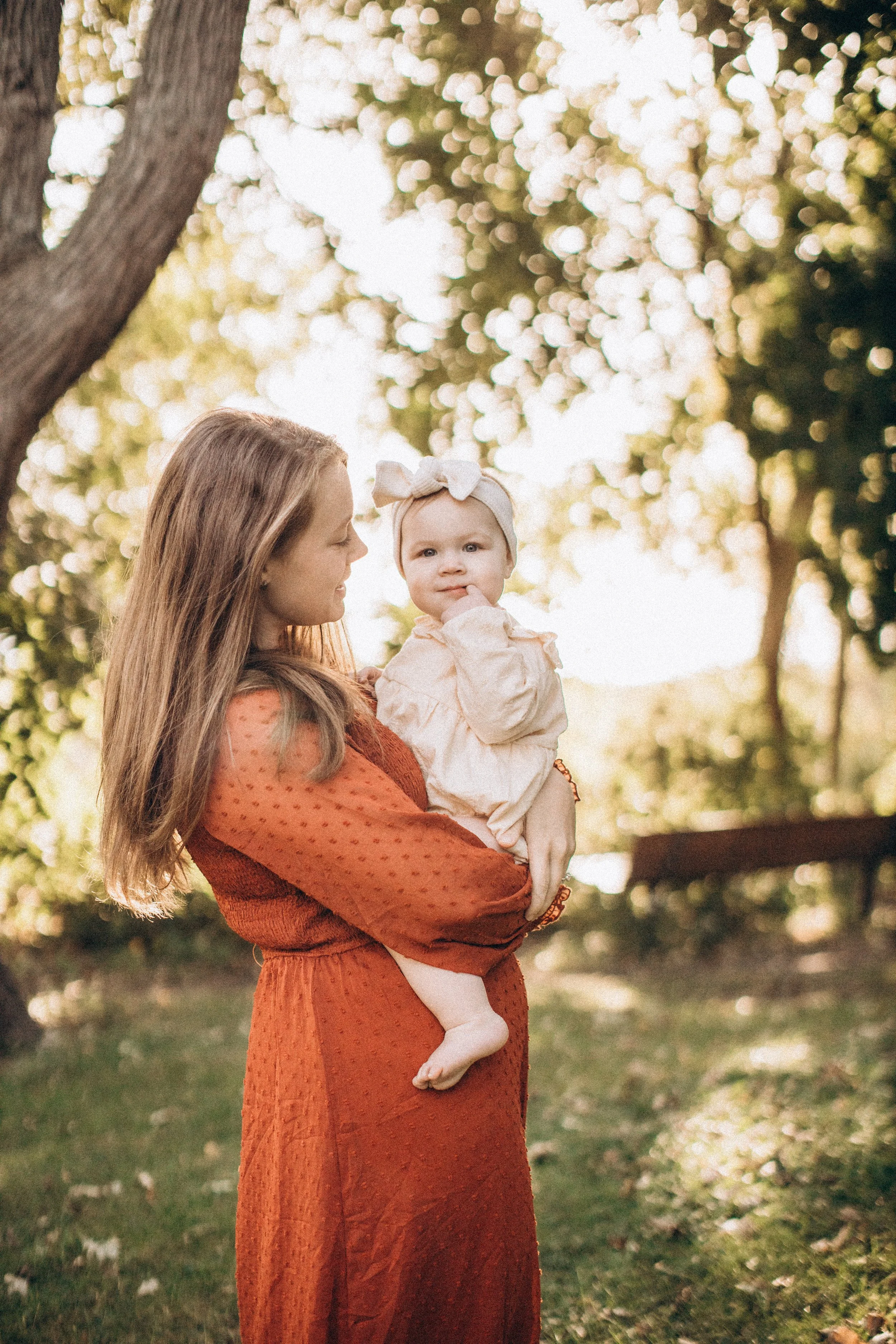 A woman with long blond hair holding a little girl outdoors in a park-like setting with trees and sunlight in the background. Lighthouse Beach Park Evanston. Family Photography