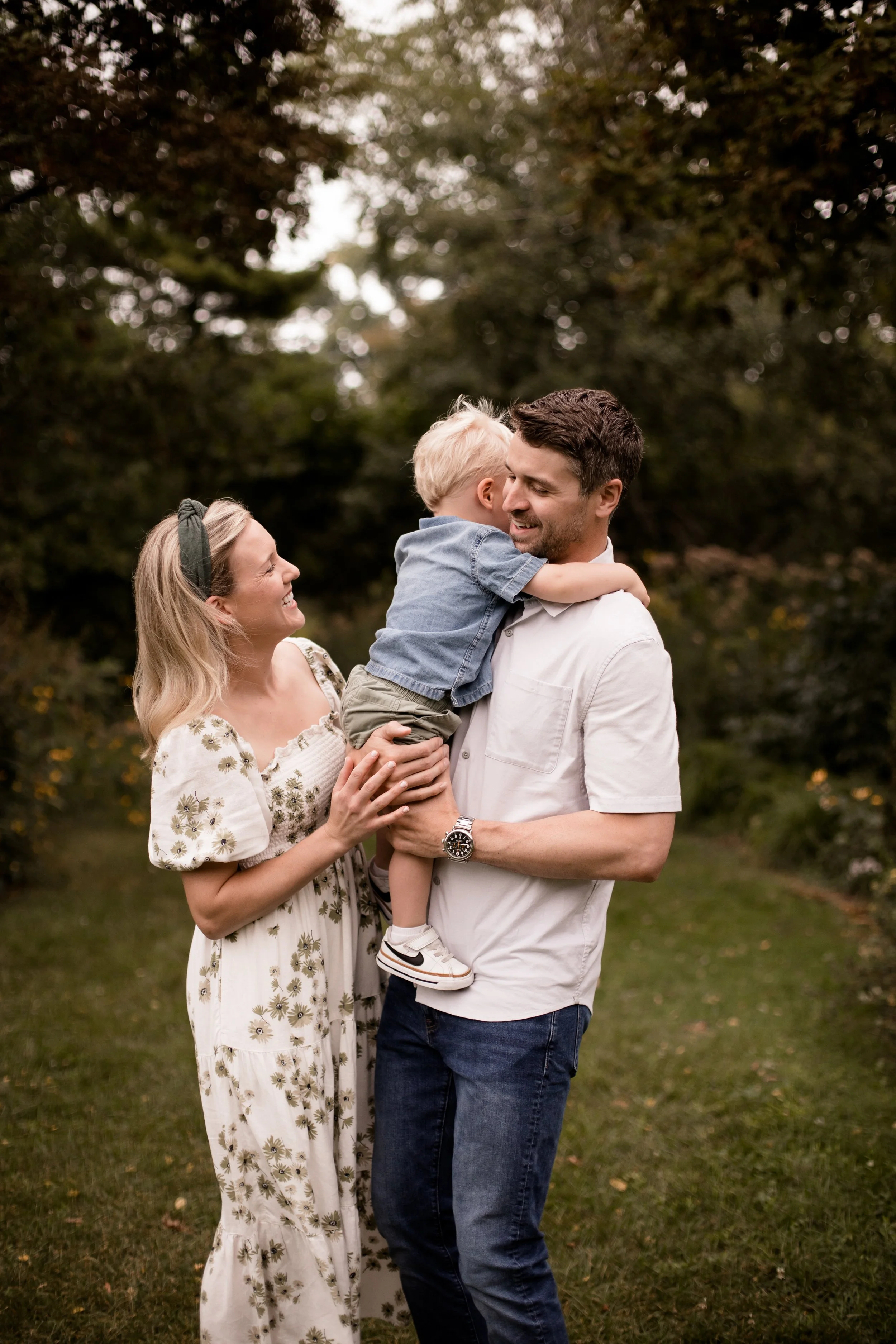 A family of three, a woman, a man, and a young boy, outdoors in a park with trees in the background. The boy is hugging the man while the woman holds his leg, and they are smiling at each other. Documentary family photography with Evanston photograph