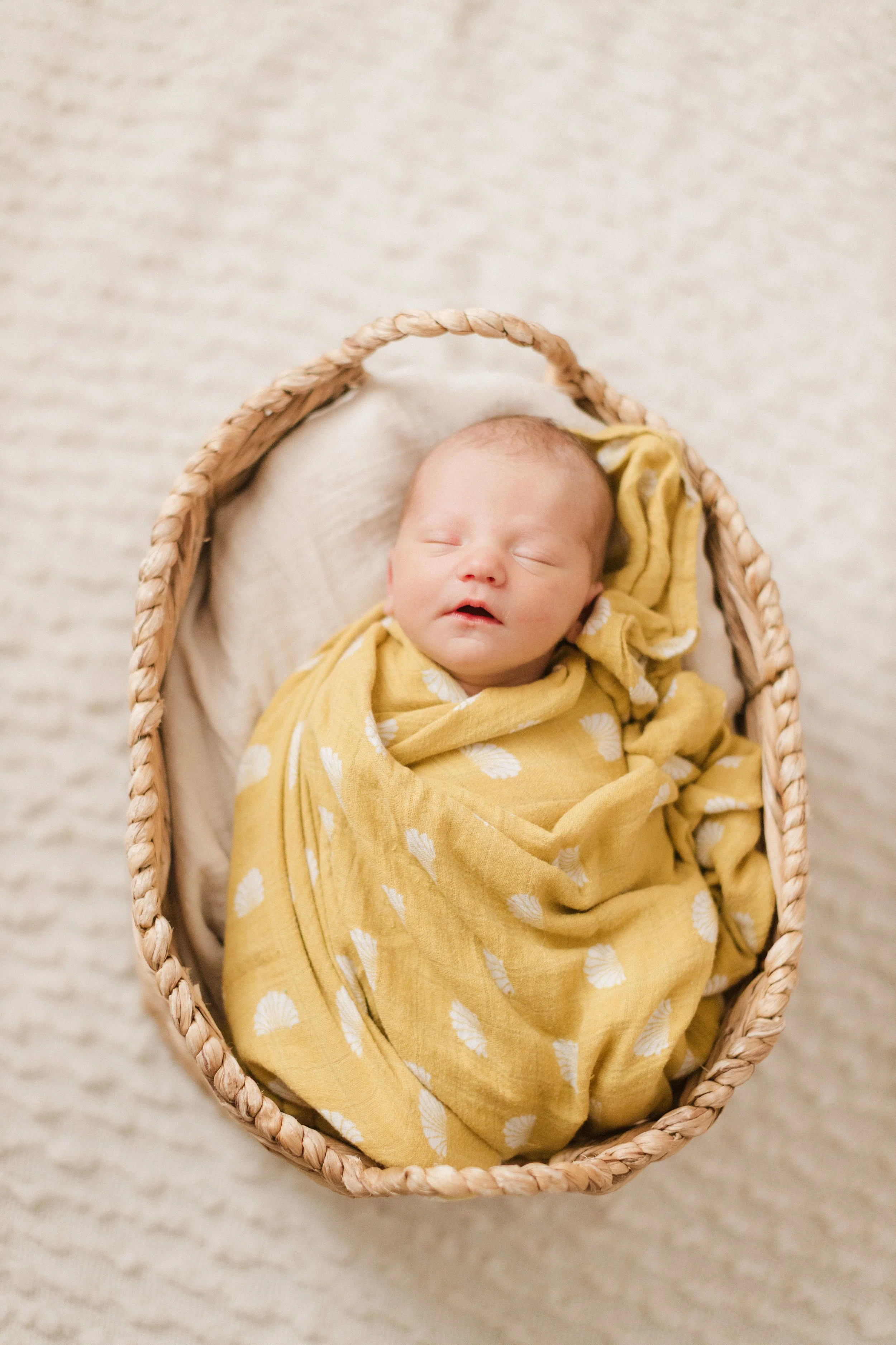 A newborn baby sleeping in a woven basket, wrapped in a yellow swaddle with white shell patterns, on a light-colored textured blanket. Lifestyle newborn session shot in Chicago IL.