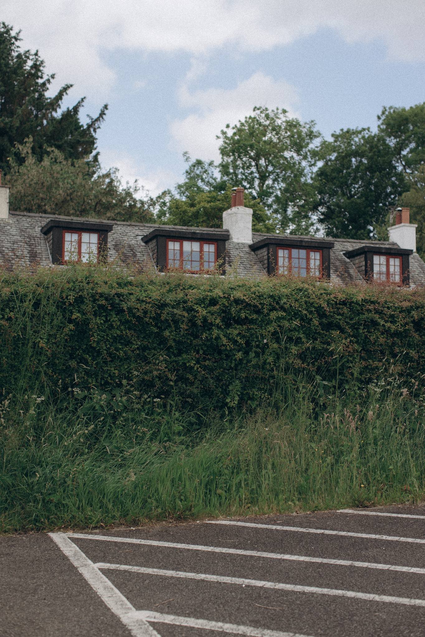 View of a house rooftop with three dormer windows, a chimney, greenery and trees, and a parking lot in the foreground.