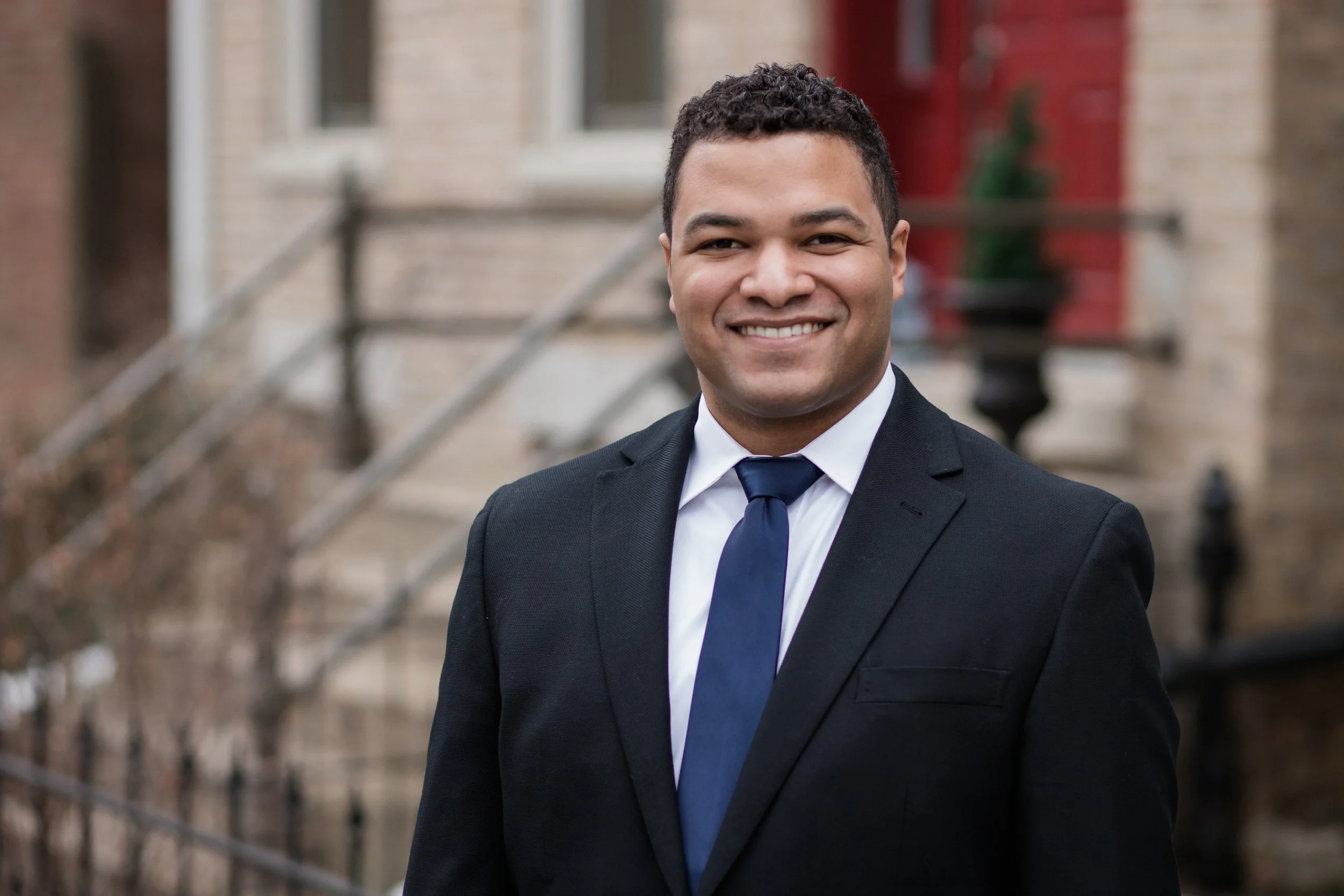 A man in a black suit with a white shirt and blue tie smiling outdoors, with a brick building and stairs in the background.