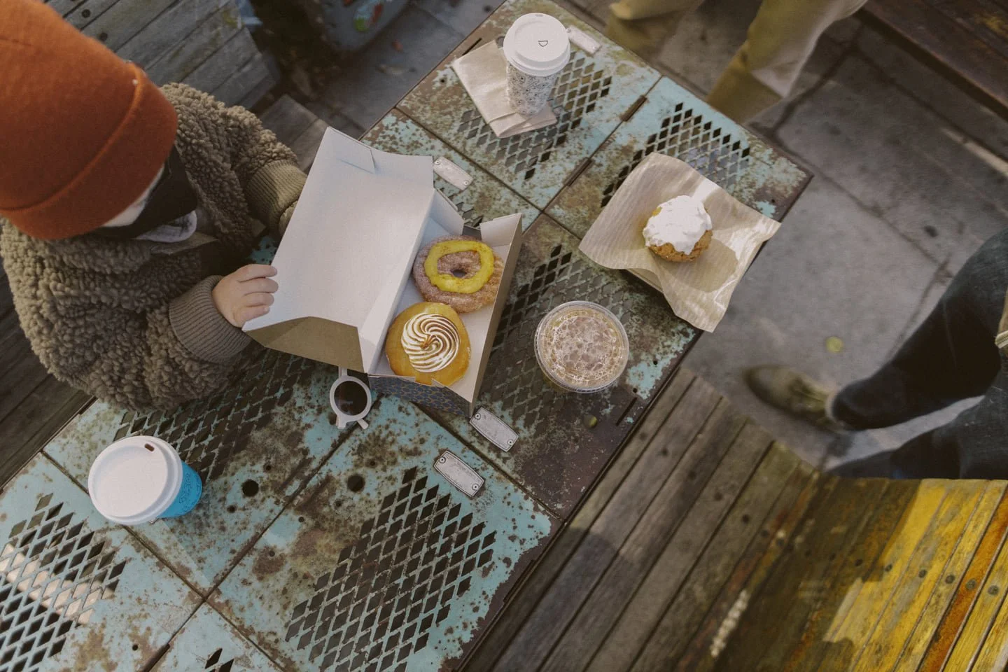 Overhead view of a rustic outdoor table with two coffee cups, a drink, and a box of assorted donuts, with two people standing nearby.