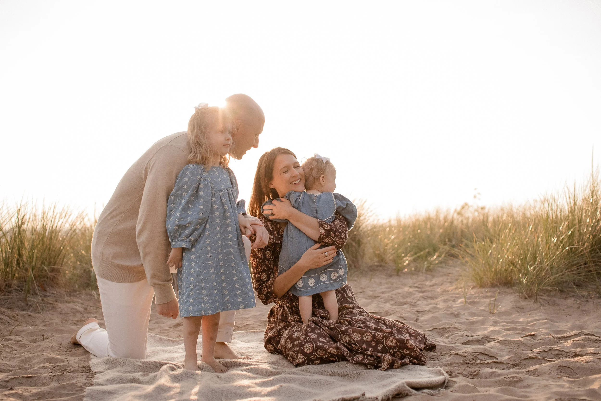 Family on the beach during sunset, with two women and two young girls, one woman is kneeling and hugging the younger girl, the other woman and girl are standing nearby, all in casual clothing, with tall grass and a bright sky in the background.
