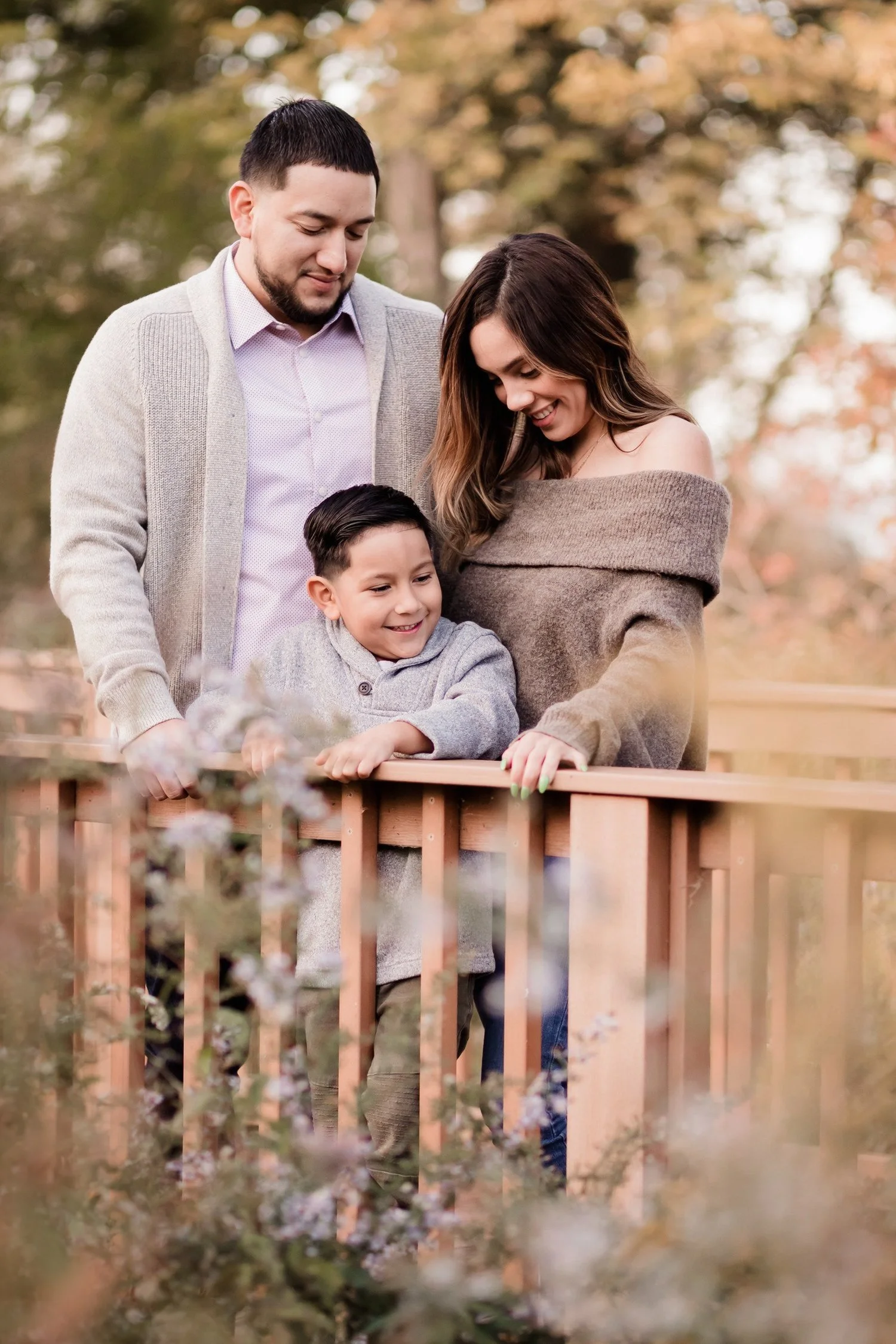 A family of three, a man, woman, and young boy, smiling and looking at something below on a wooden railing outside during autumn, surrounded by colorful fall foliage. Gillson Park, Wilmette. Evanston based photographer