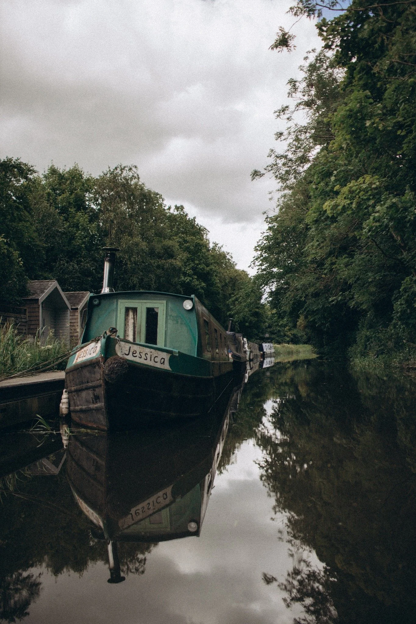 Black and green narrowboat named Jessica moored along a calm canal with dense green trees and cloudy sky reflection in the water.