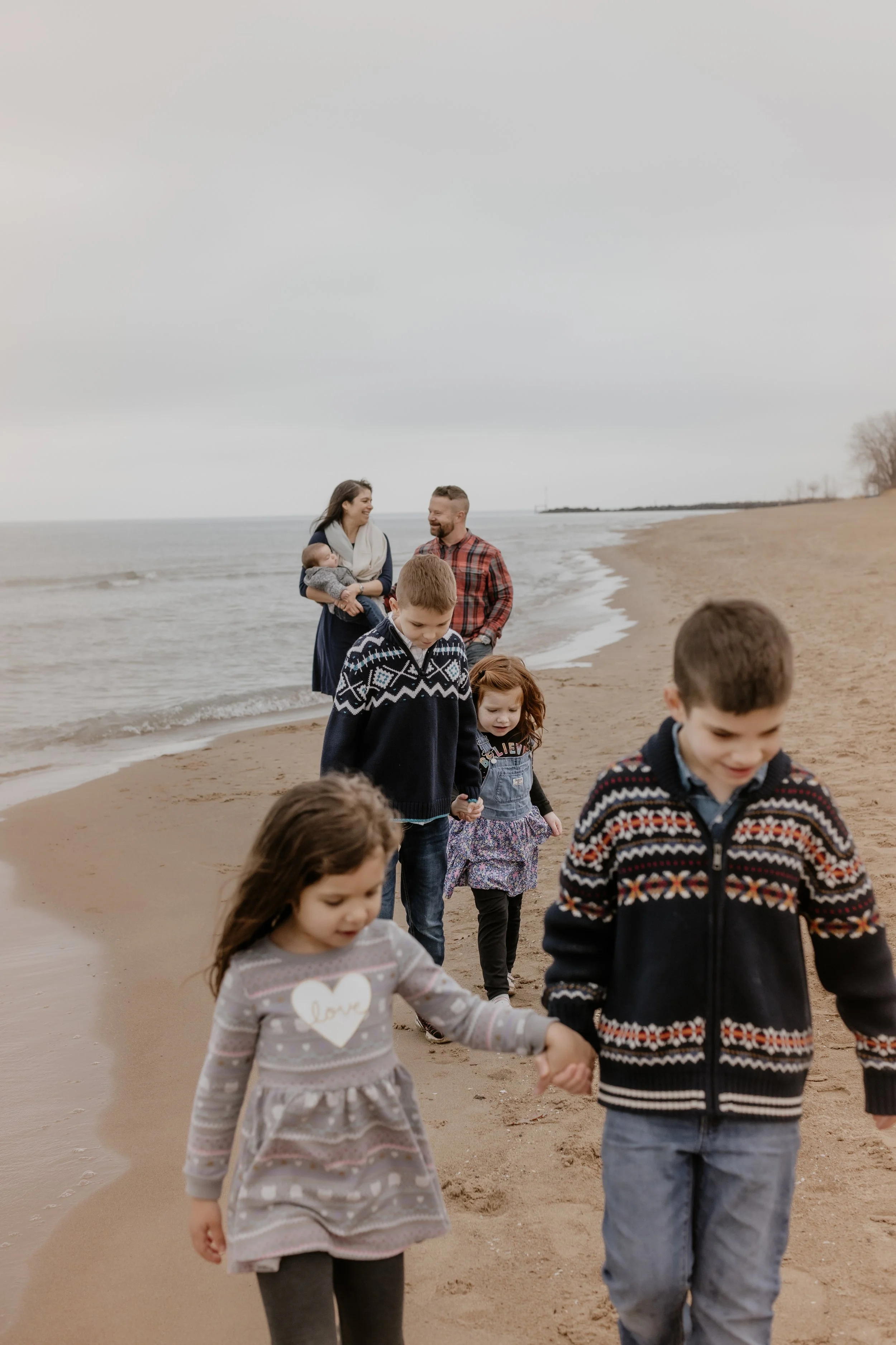 Family walking along the beach, holding hands, with a woman and man in the background carrying a baby in their arms.