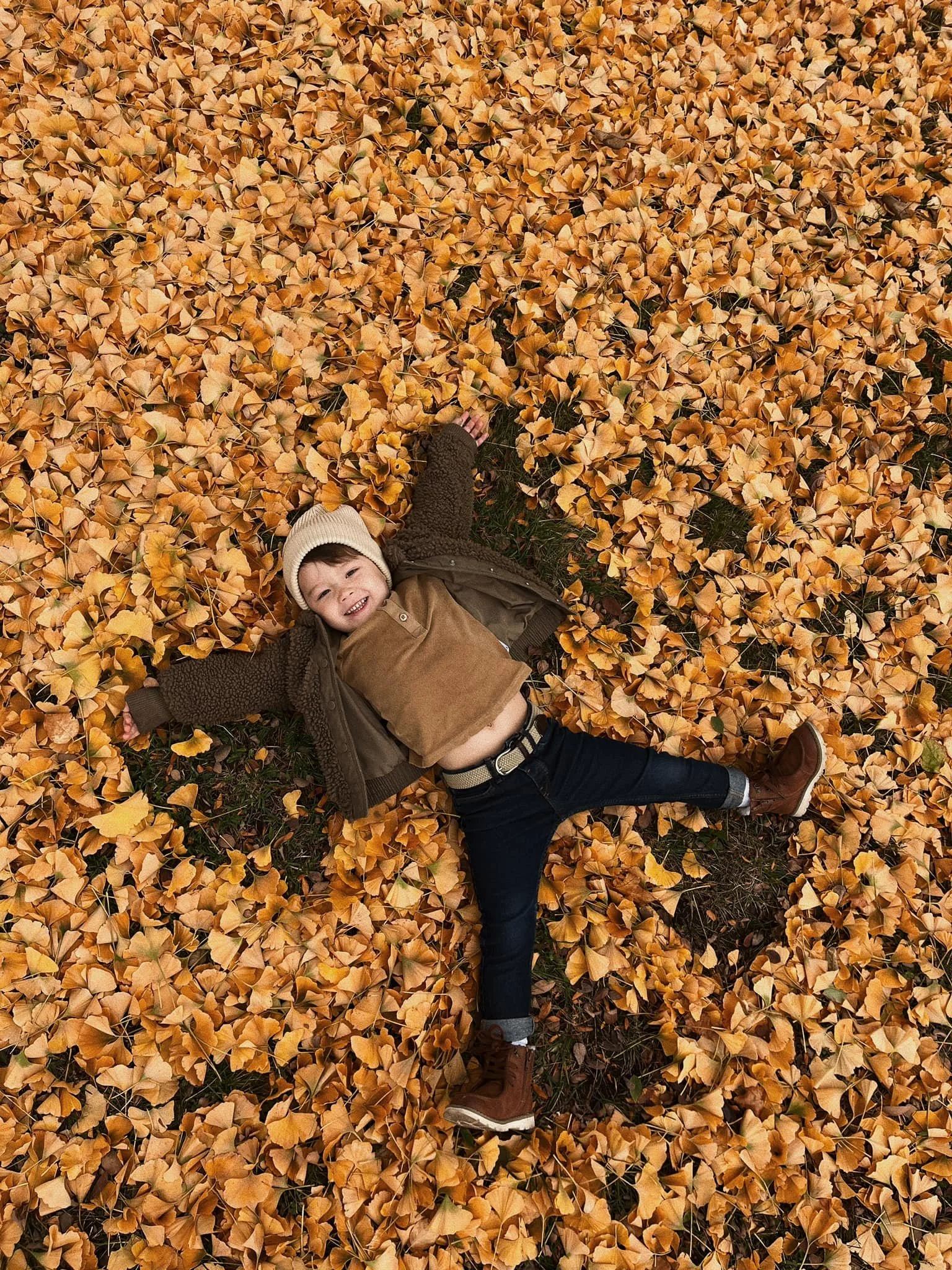 A young boy lying on the ground covered with golden autumn leaves, wearing a beige beanie, brown jacket, dark jeans, and brown boots, smiling and looking up.