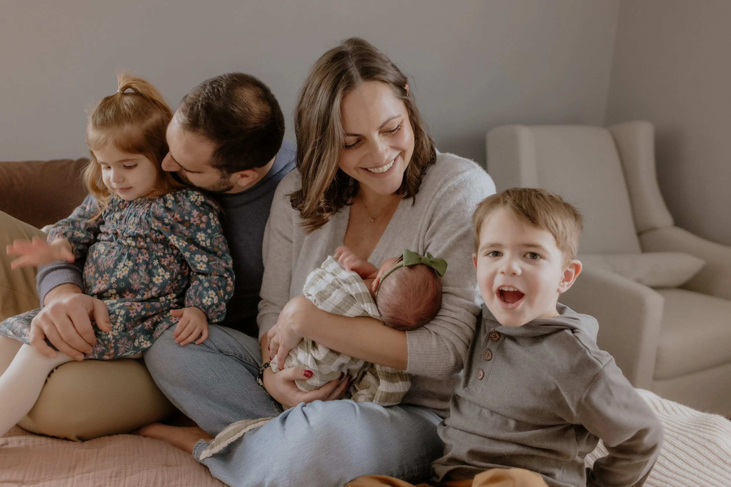 Family sitting on a couch, smiling, with a woman holding a newborn baby, a young girl leaning on a man's lap, and a boy making a playful expression. Lifestyle newborn session shot in Evanston IL.