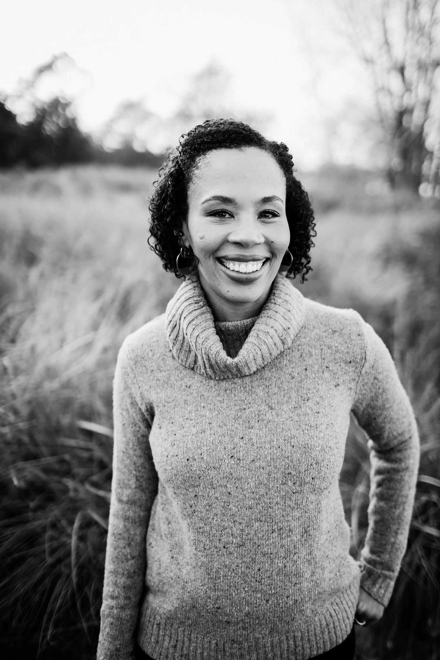 Black and white photo of a woman smiling outdoors in a natural setting, wearing a cozy turtleneck sweater.