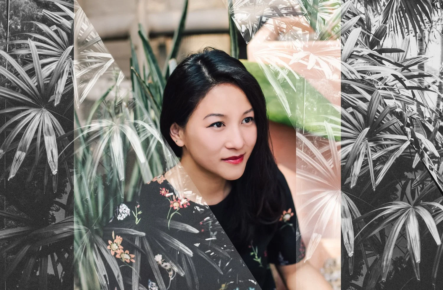 A woman with long black hair, wearing a black floral top, sitting among green and black-and-white tropical plants, smiling gently. Headshot photography session in Evanston, IL.