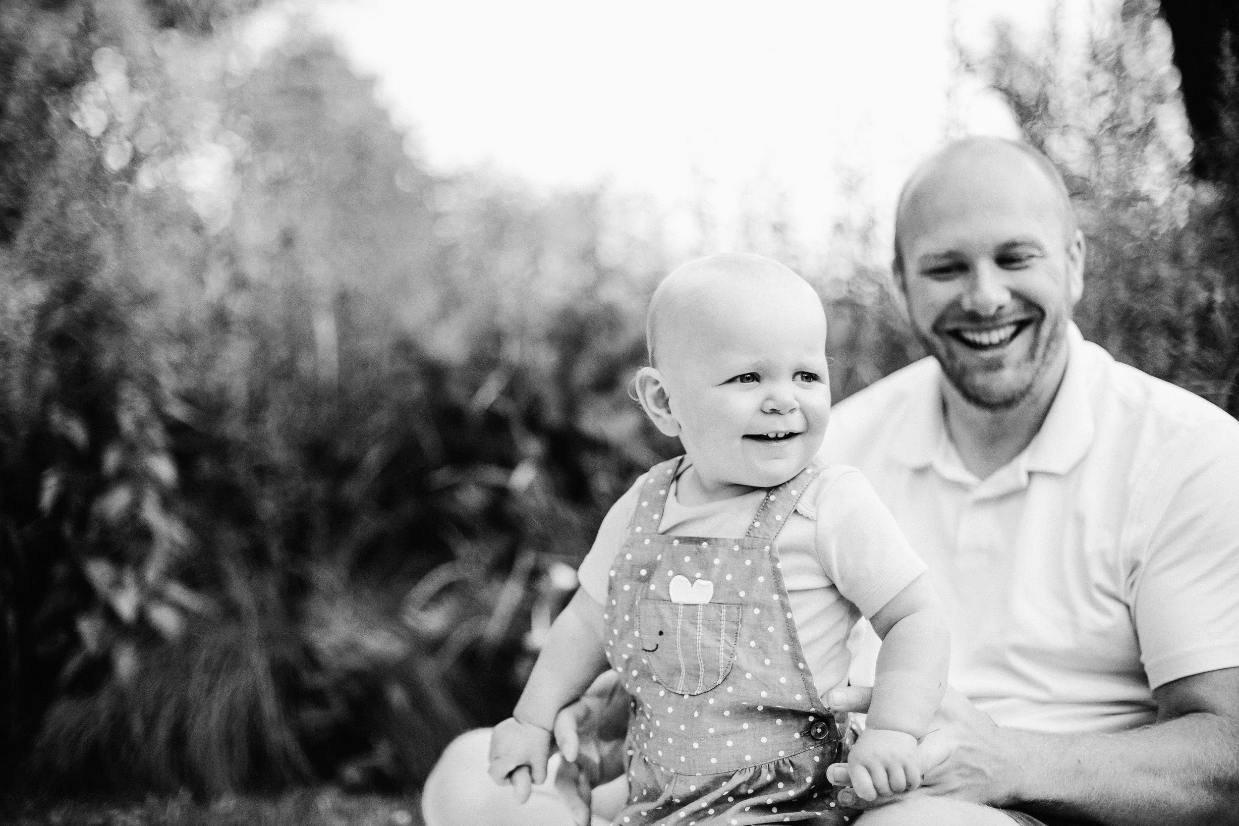 A smiling man and baby girl sitting outdoors, the man holding the baby in his lap, both dressed casually, with trees and natural background. Family photographer Evanston IL.