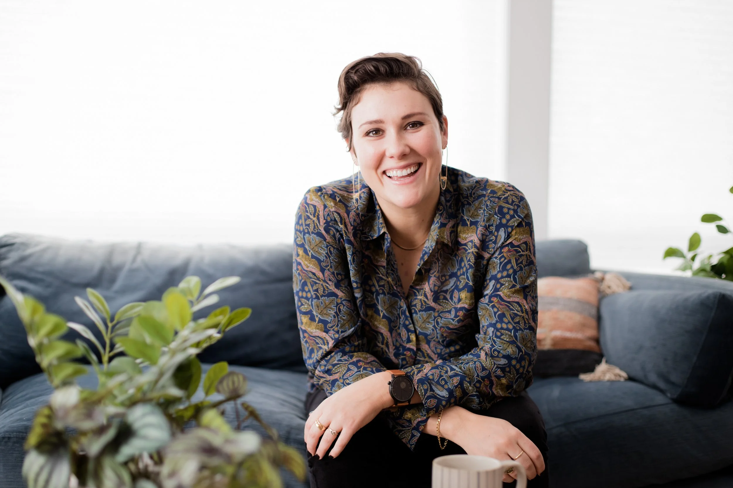 A smiling young woman with short brown hair, wearing a patterned blouse, sitting on a dark blue sofa in a bright room, with a plant in the foreground and a coffee mug in her hand.