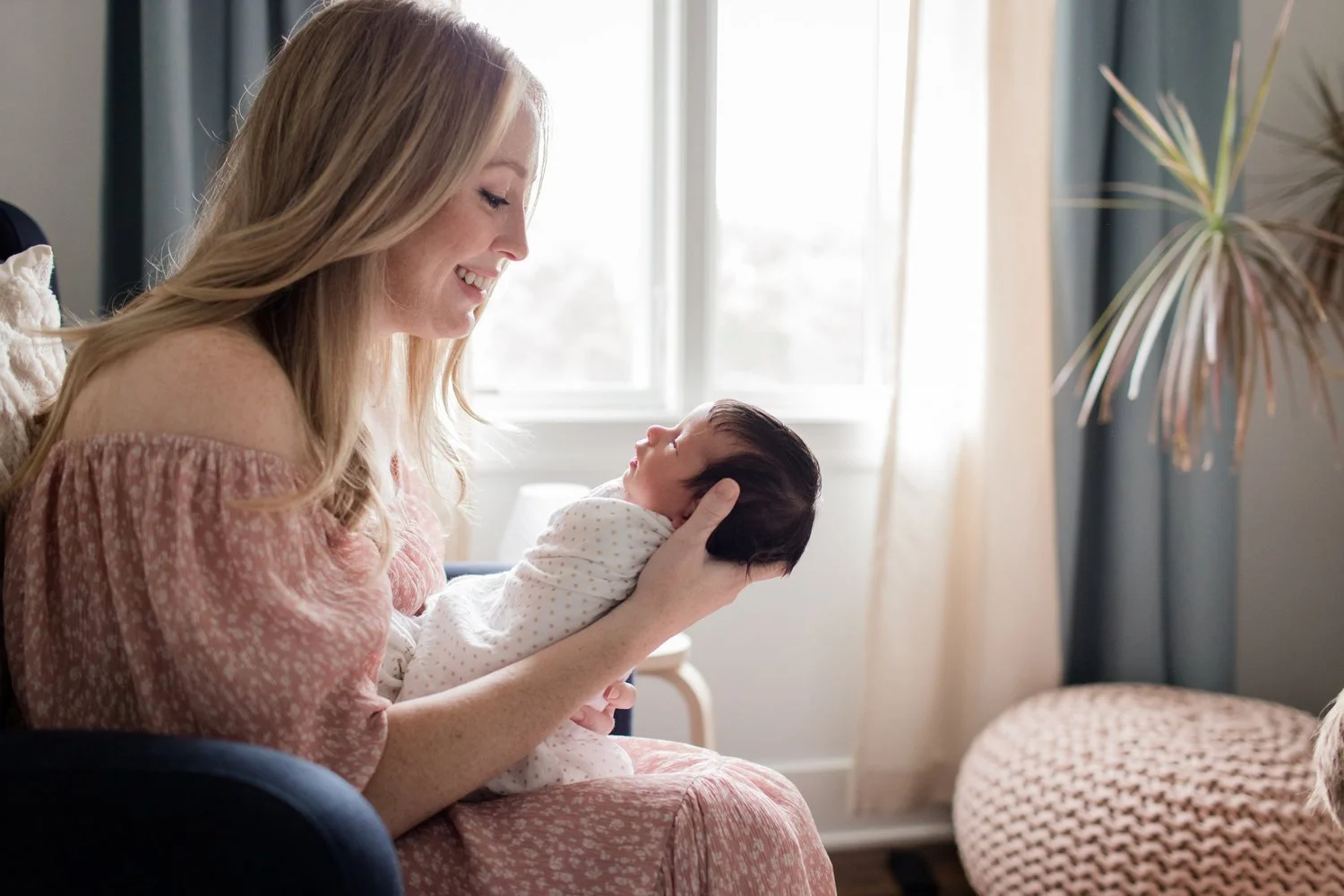A woman holding a newborn baby indoors near a window with sunlight. She is smiling and looking at the baby, who is wrapped in a white blanket with small polka dots.
