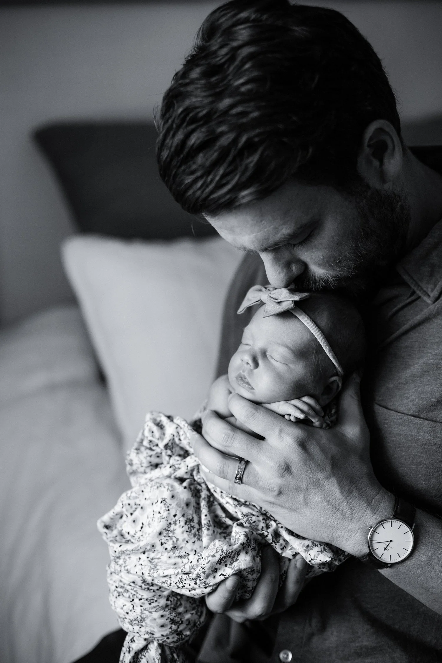 A man with dark hair and a beard holding a sleeping newborn girl with a headband and floral dress.
