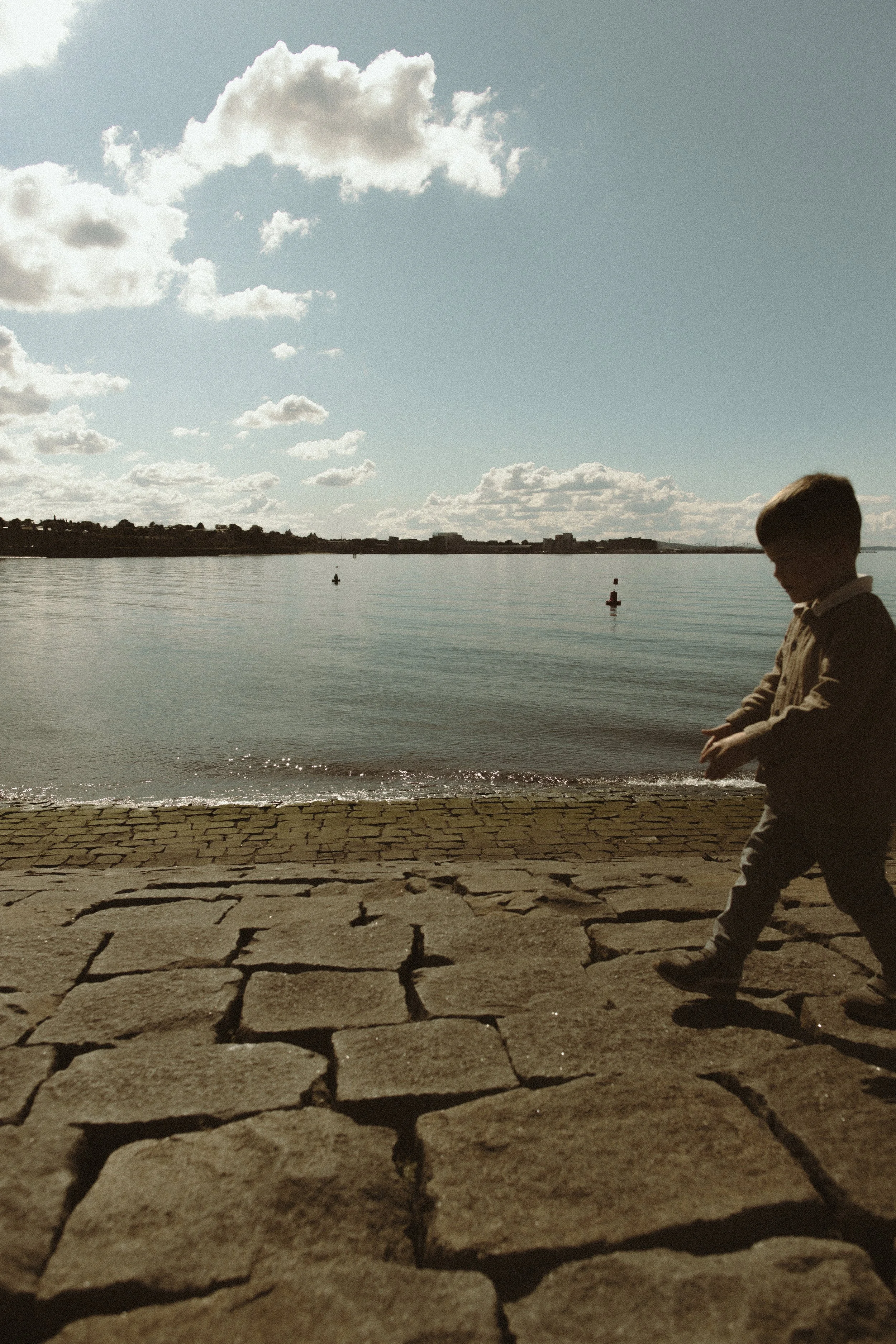 A young boy walking along a stone-paved waterfront under a partly cloudy sky with calm water in the background.