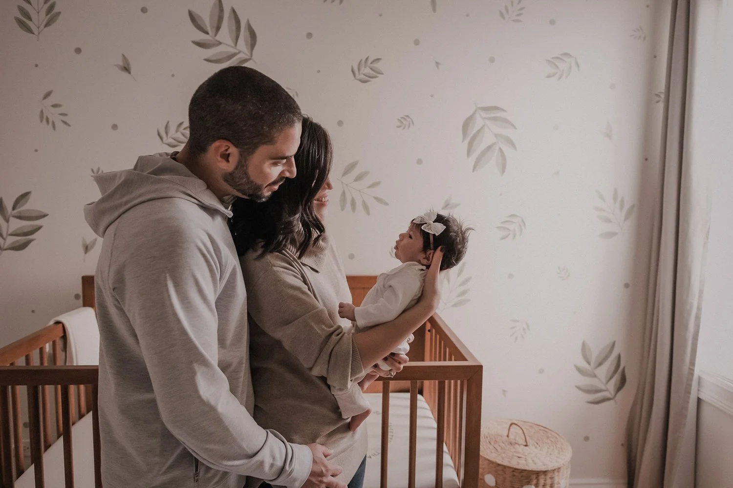 A family of three in a nursery, with a man and woman looking at their infant daughter, who has a bow in her hair, inside a crib. Newborn photography in Chicago IL. Focusing on a candid storytelling approach.  