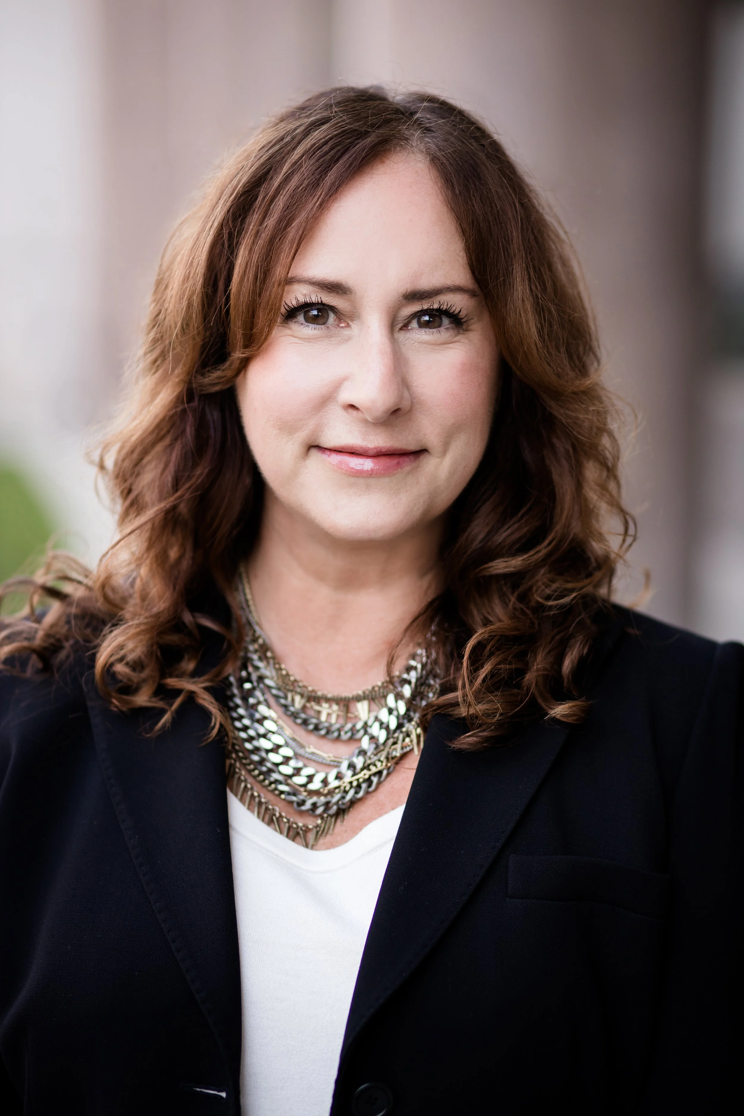 Headshot of a woman with shoulder-length wavy brown hair, wearing a black blazer, white shirt, and layered silver necklaces, smiling outdoors. Headshot photography session in Evanston, IL.