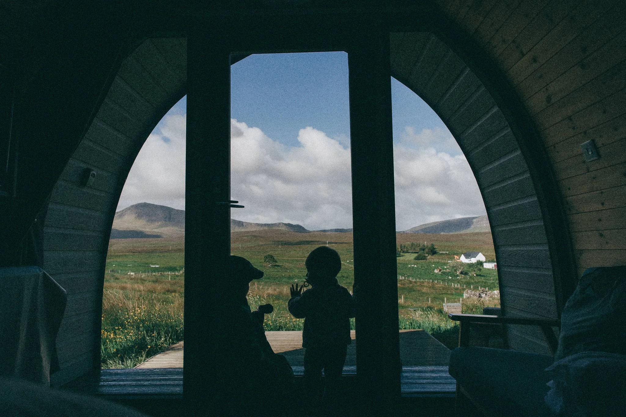 Silhouettes of two children looking out a door to a field with mountains in the distance, inside a wooden house or barn.