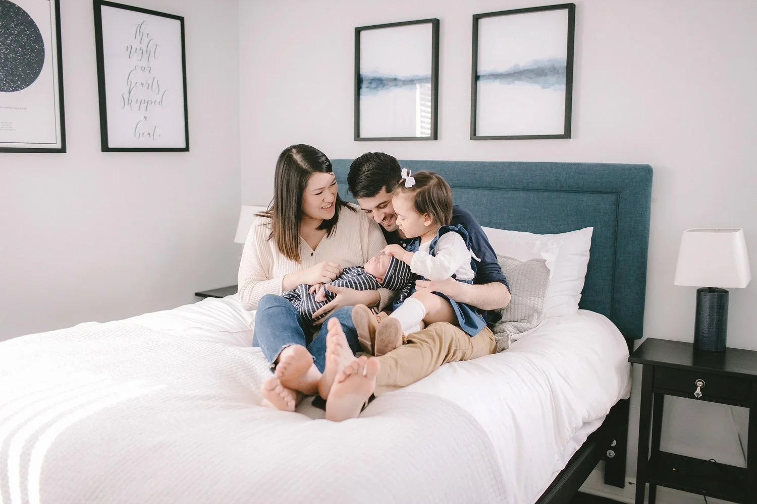 A family of four sitting on a bed, smiling and looking at a newborn baby. The mother, father, and young girl are all close together, embracing the baby. The bedroom has framed artwork on the wall and a nightstand with a lamp. Chicago IL newborn