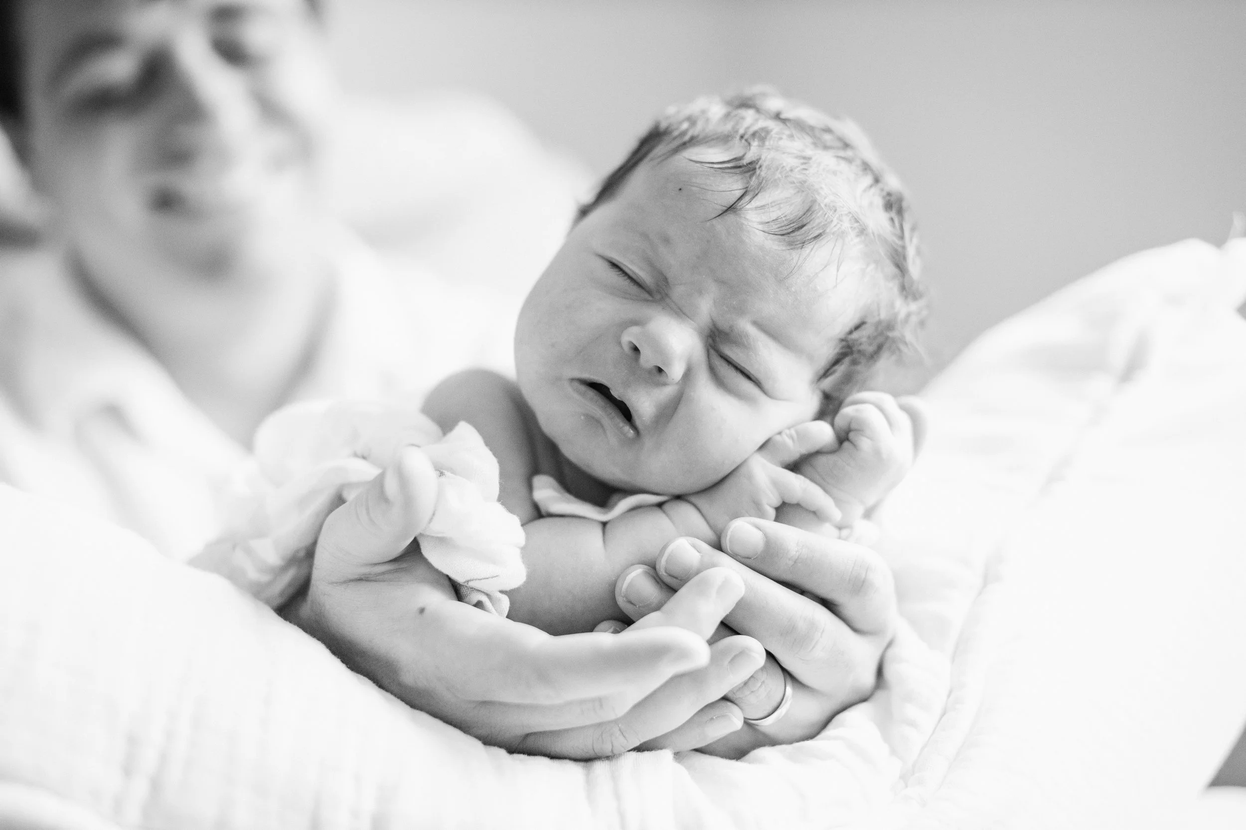 A newborn baby being held by a person, with the baby's eyes closed and mouth slightly open, in black and white.