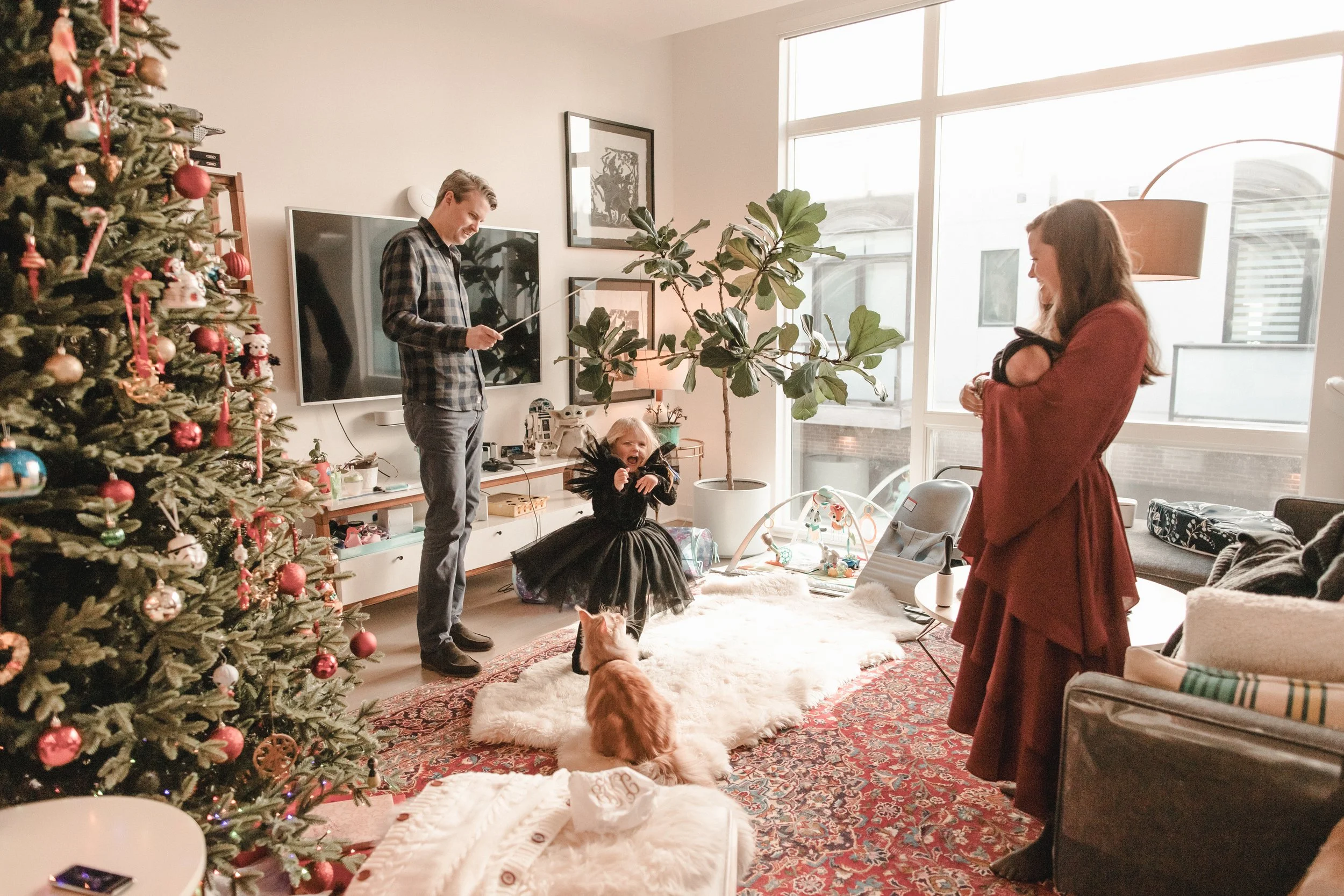 Family celebrating Christmas in living room with decorated tree, child in black dress, woman in red dress holding a cat, man in plaid shirt watching, dog on fluffy rug, large window in background. Lifestyle newborn session in Chicago IL.