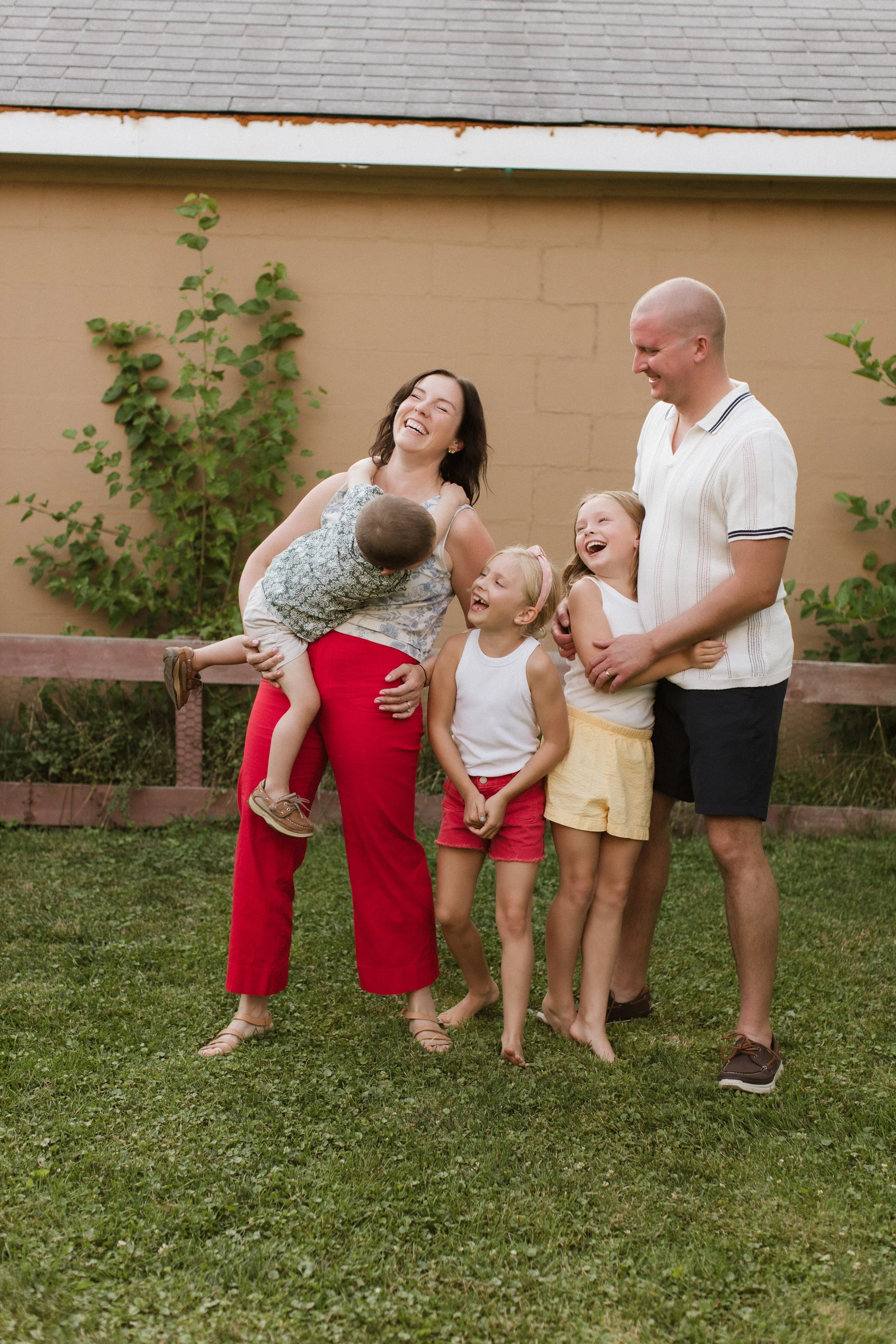 A family of five laughing and playing outdoors in a grassy yard with a beige wall and green foliage in the background. The mother is holding a young boy, and the father is embracing a girl; two other girls stand nearby.